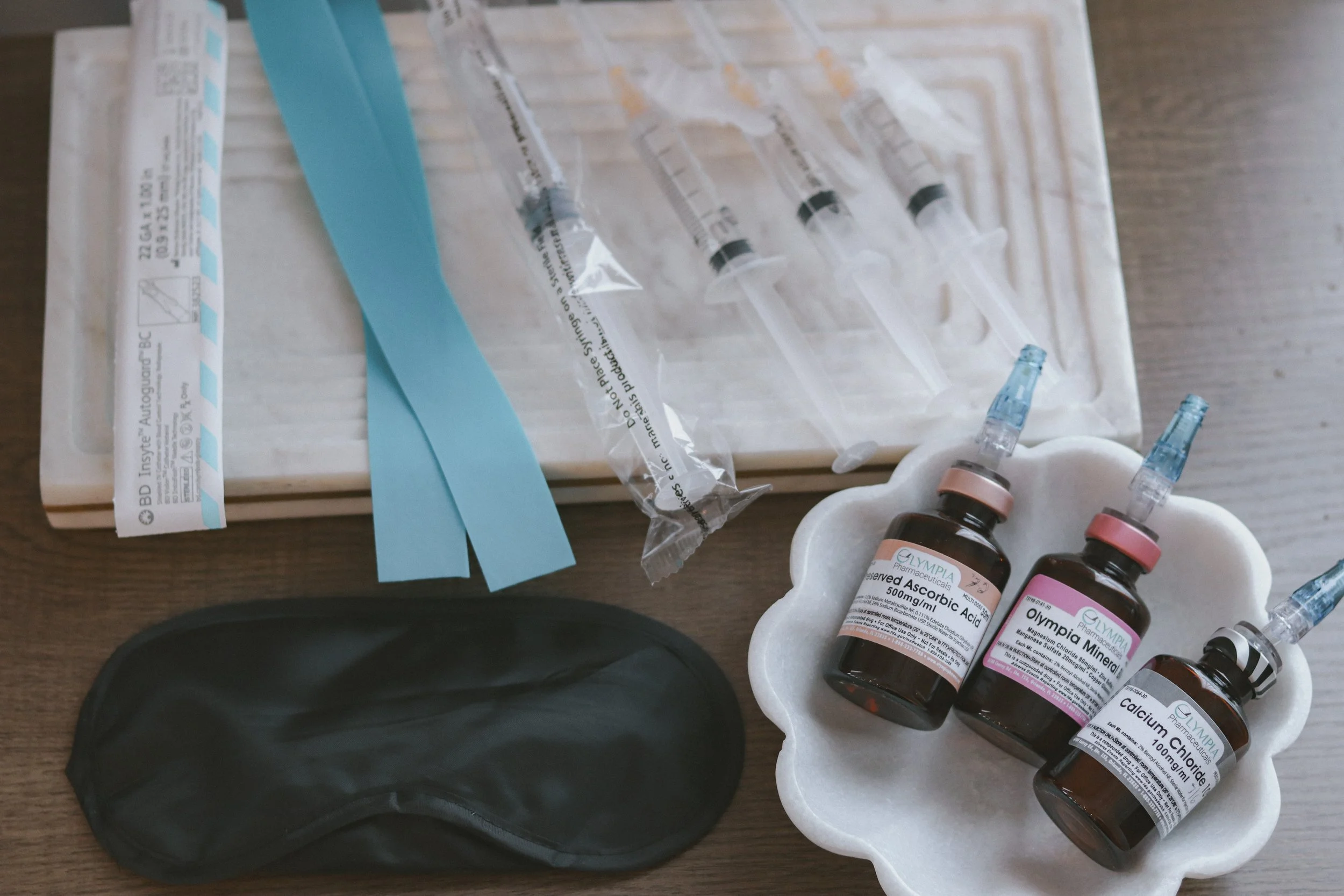 Medical supplies including syringes with needles, bottles of preserved ascobic acid, Olympia mineral, calcium chloride, a black eye mask, and packaging on a wooden surface.