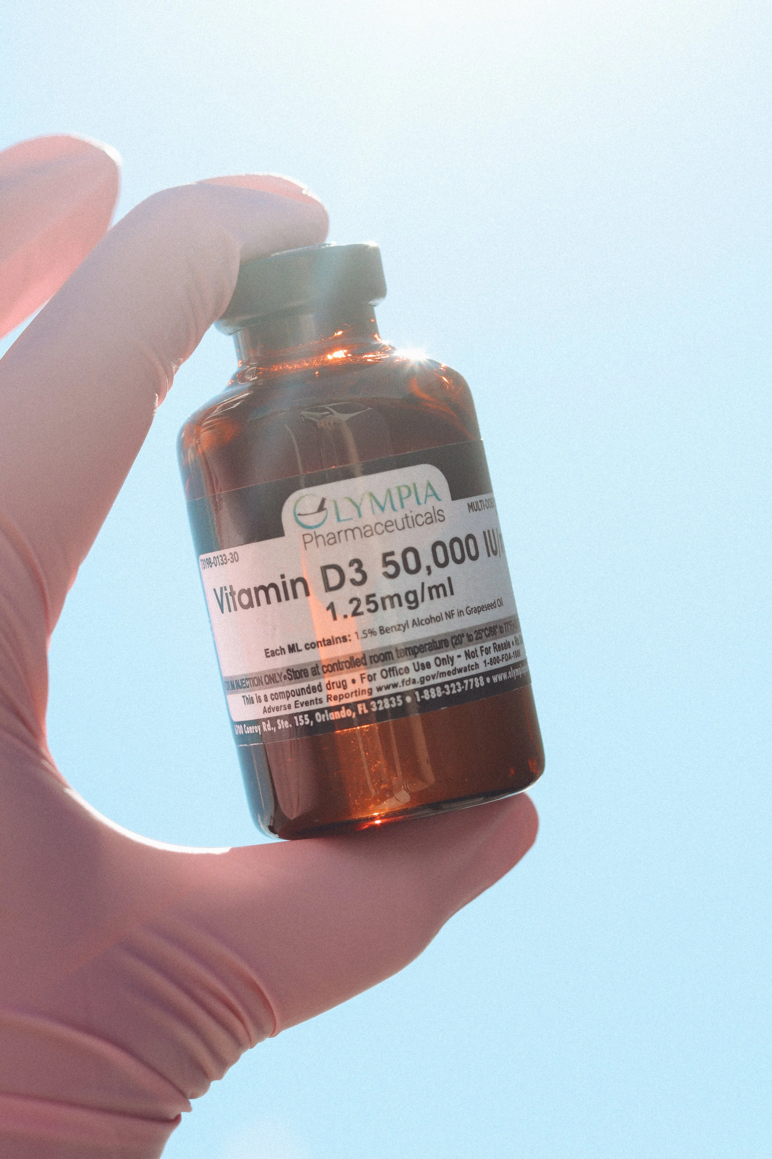 A hand wearing a pink glove holding a small brown glass bottle labeled 'Vitamin D3 50,000 IU 1.25 mcg/ml' against a light blue sky background.