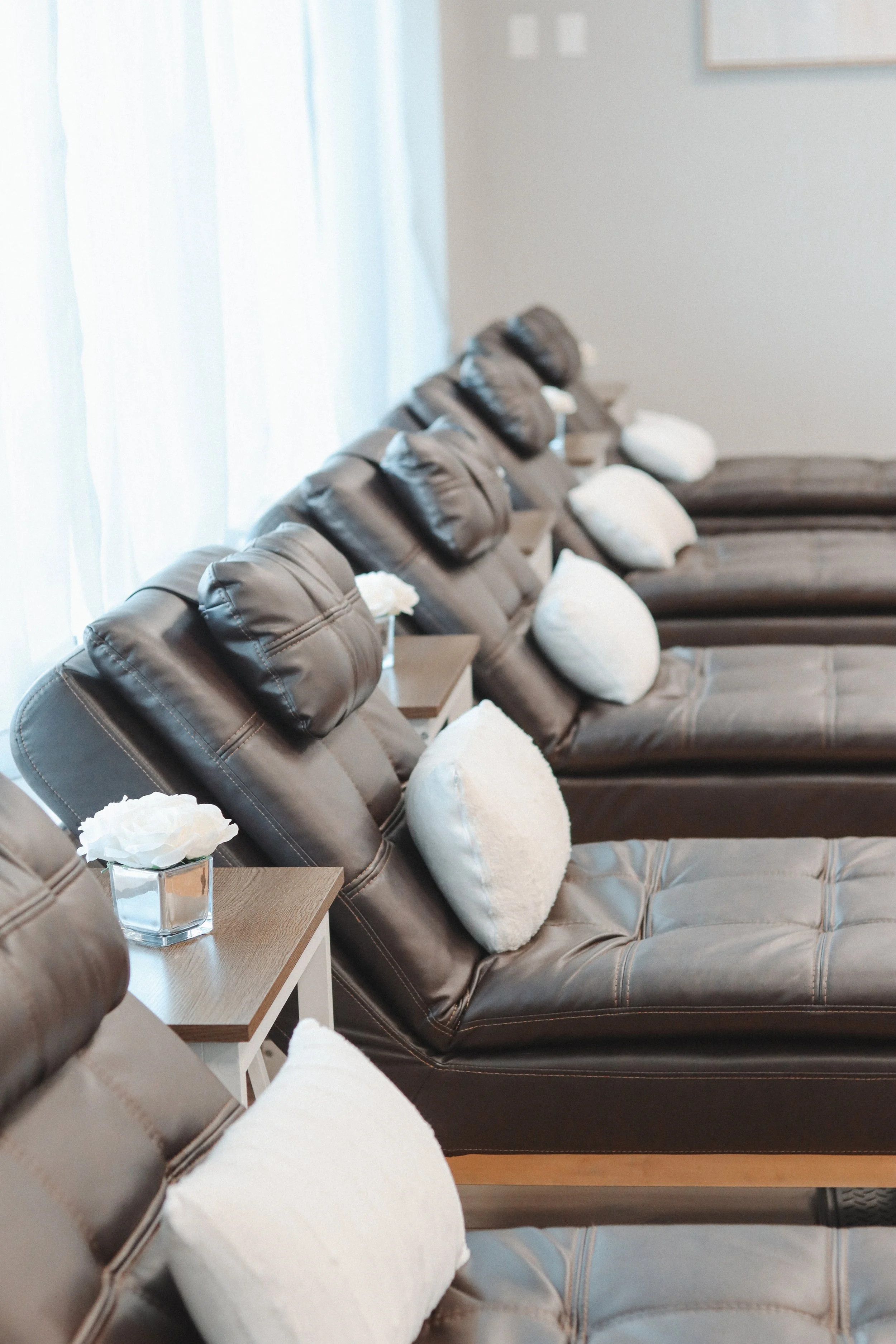 A row of brown leather recliners with white pillows, small side tables with white flowers, and sheer white curtains in a waiting area.