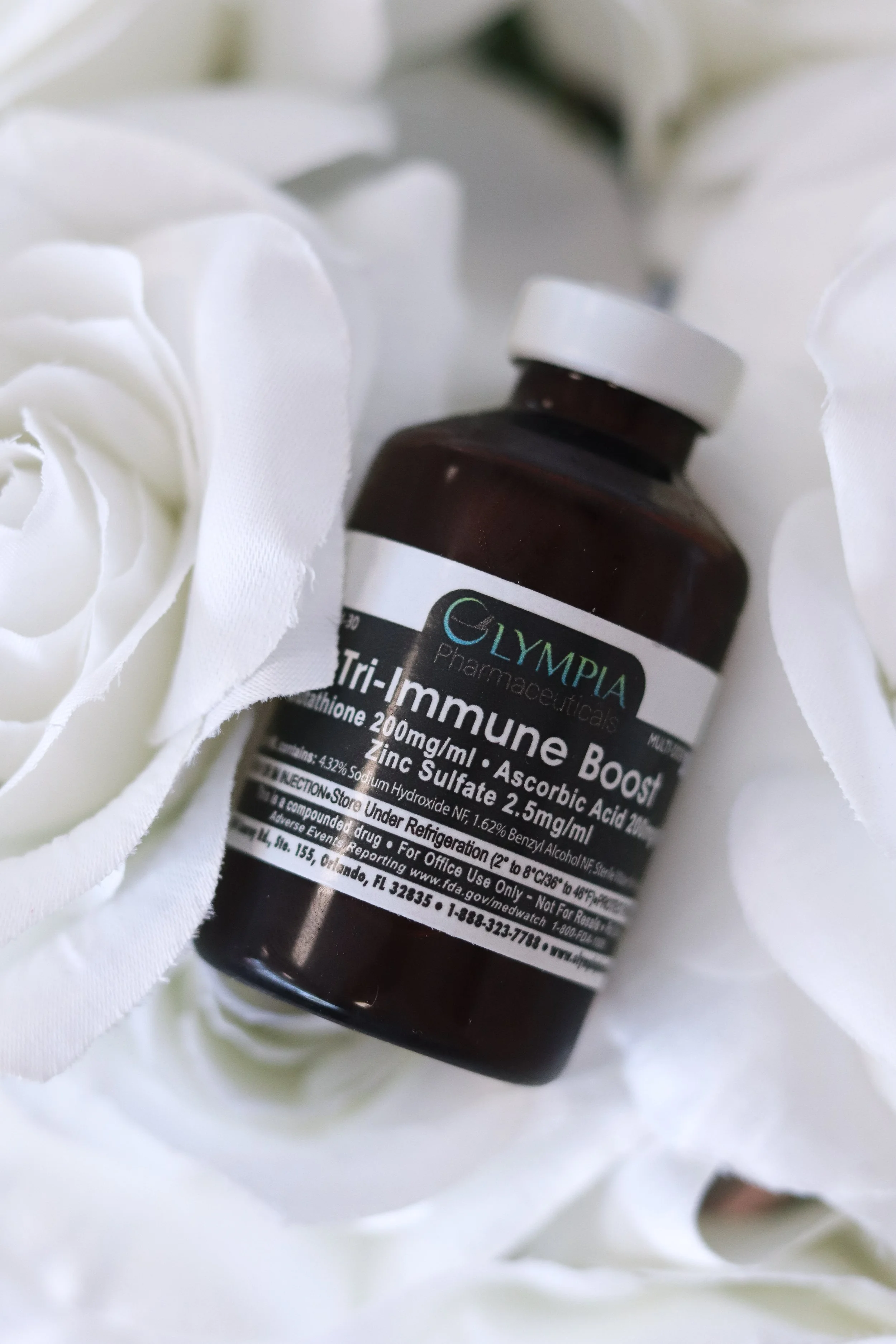 A brown bottle of Olympiad Tri-Immune Boost supplements lying on white fabric with roses.