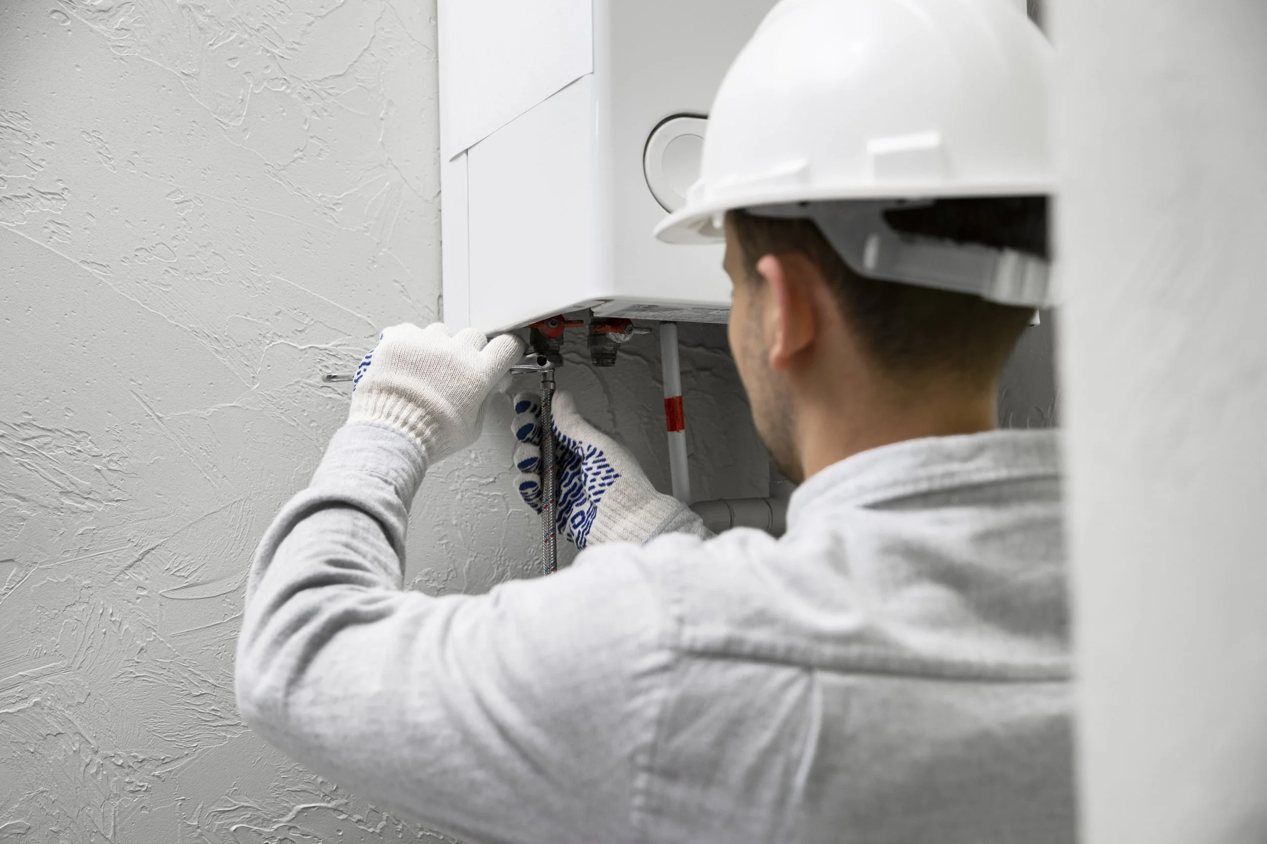 Close-up of a heating engineer repairing a home boiler system, tools in hand, with pipes and controls visible against a clean domestic background.
