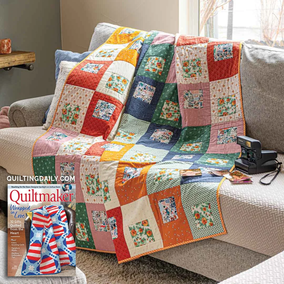 The Snapshot quilt draped casually over a gray sofa in a warm, natural-light living space, showcasing the quilt's vibrant mix of floral novelty prints framed by bold solid blocks in red, navy, teal, gold, pink and cream.