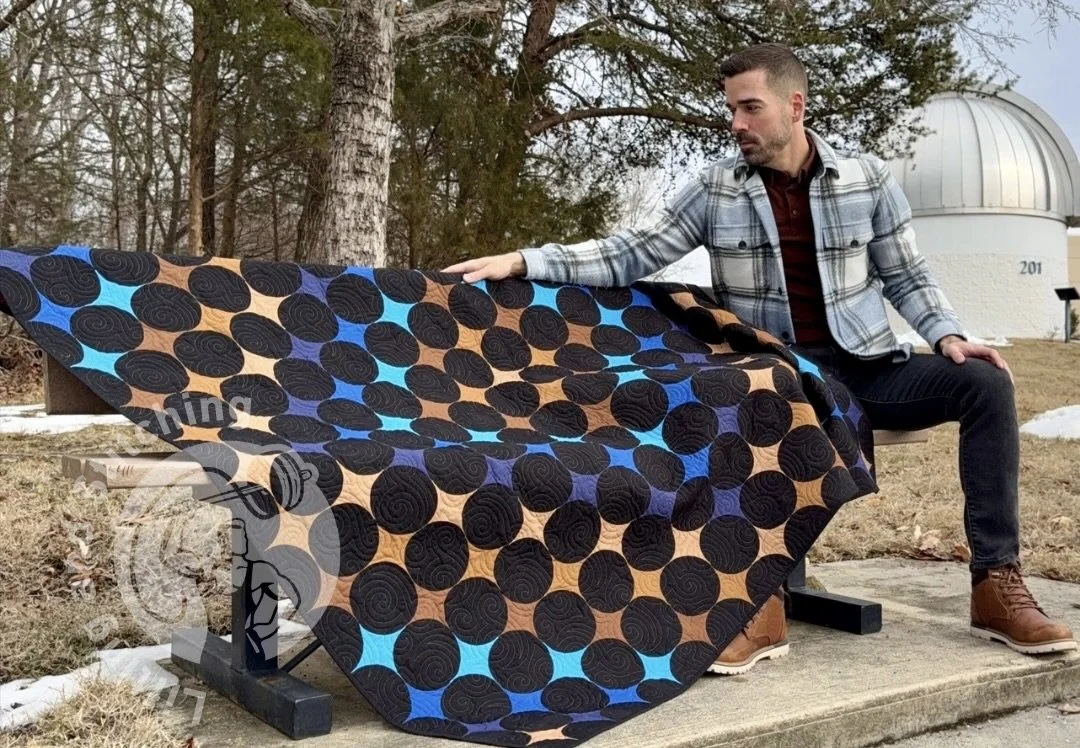 A person sits on a wooden bench outdoors with a large modern quilt featuring four‑point stars in browns and blues and an all‑over swirling motif. Winter trees, patches of snow, and a space observatory dome appear in the background.