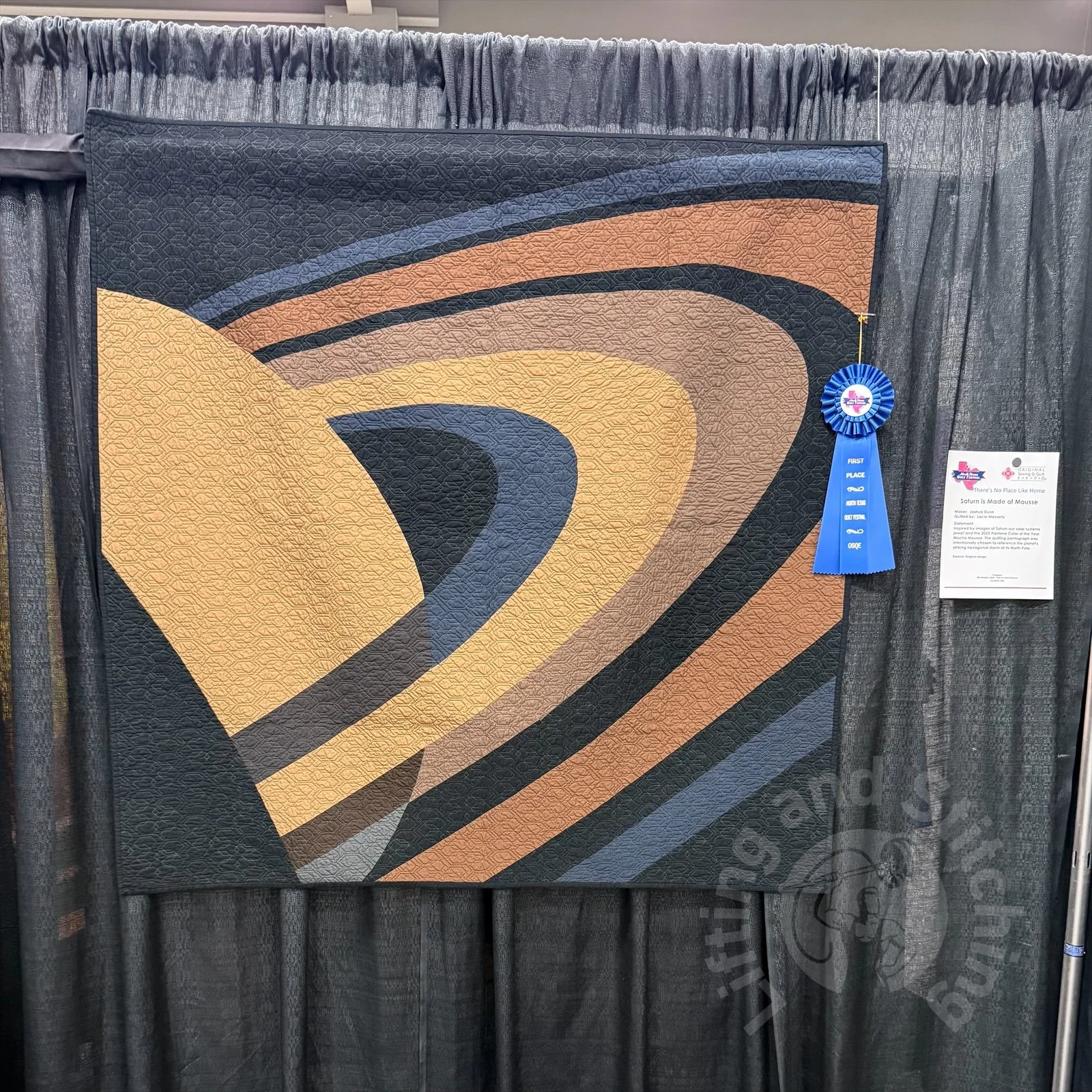 A quilt of Saturn on exhibit the North Texas Quilt Festival adorned with a blue ribbon, winning first place in the modern category.