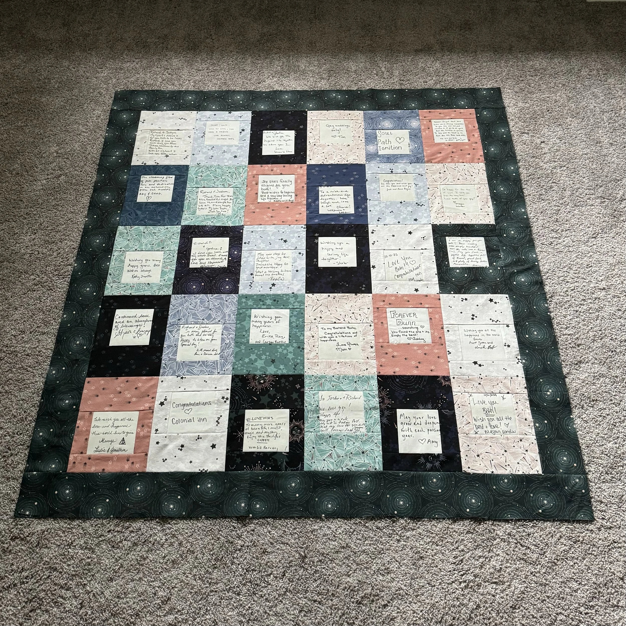 A completed wedding guest signature quilt in celestial navy, teal, blush and grey prints, laid flat on carpet.