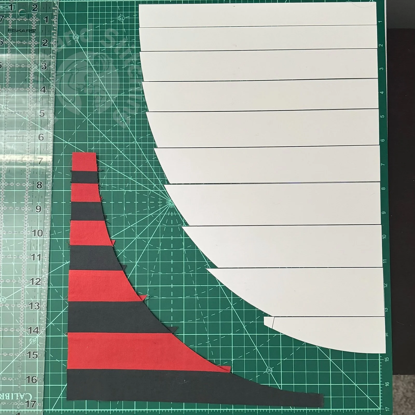 A flat lay image of fabrics strips pieced together in alternating red and black colors on a cutting board. On the other side is a set of cardboard templates.