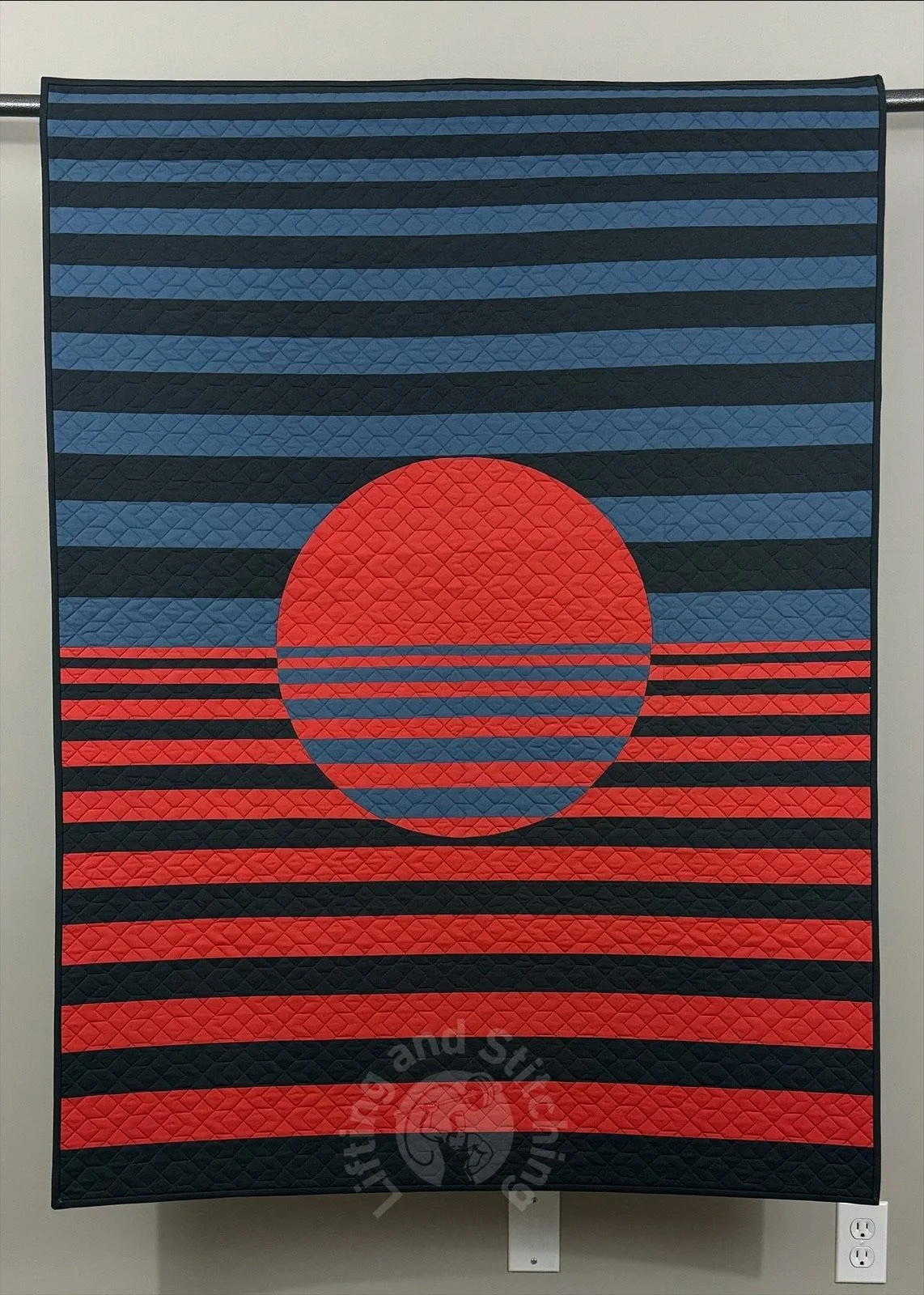 A wall hanging quilt hanging on a rode for its photograph. The quilt appears straight on to the frame with a giant circular red center, half in stripes. The top and bottom half to the quilt around the circle are striped horizontally.