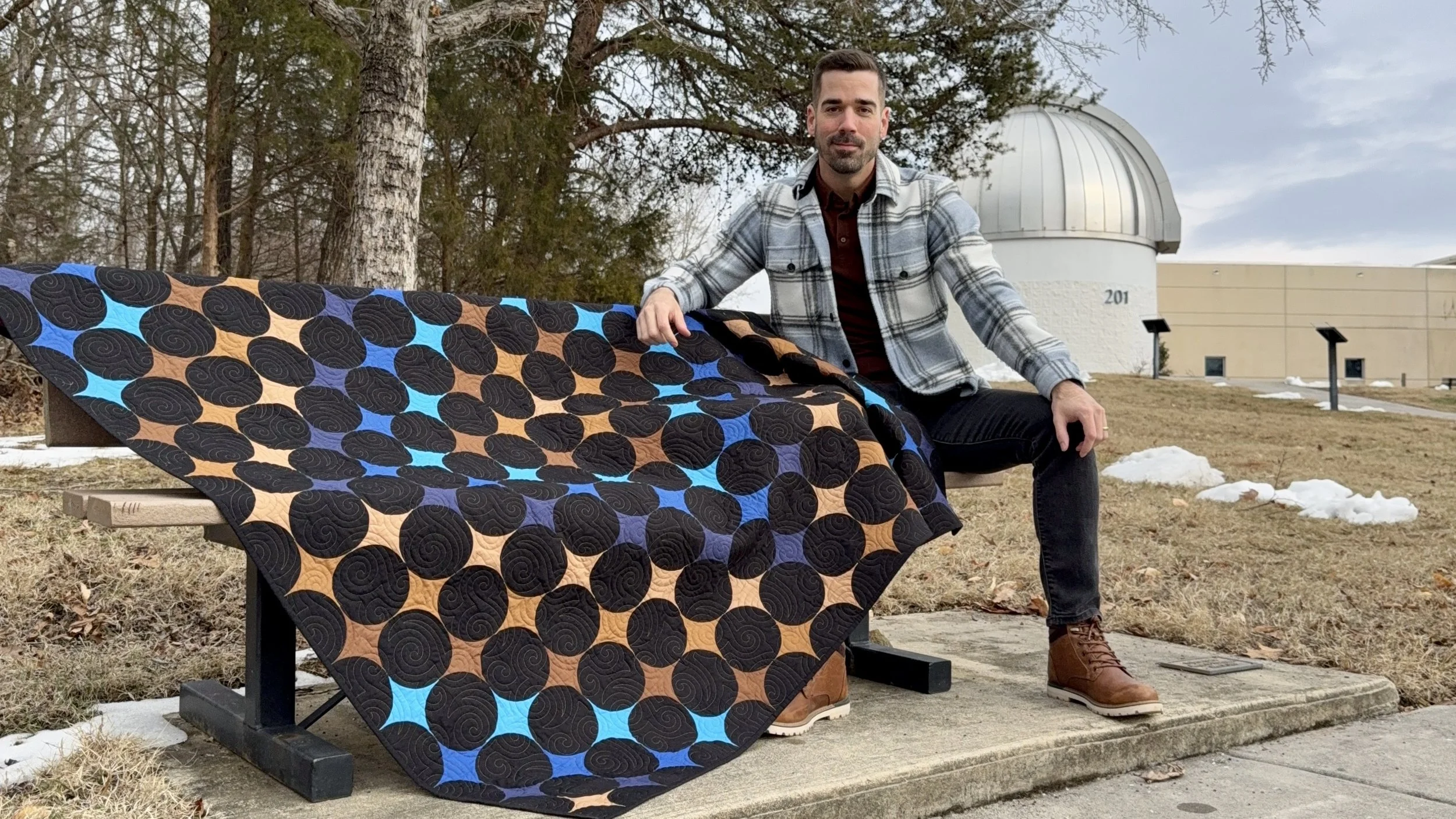 Quilt titled A Trip Around the Universe with a flowing wave of brown and blue fabric arranged into retro-modern four-point stars, the designer Joshua Dunn of Lifting and Stitching sitting beside it on a park bench with an observatory in view.