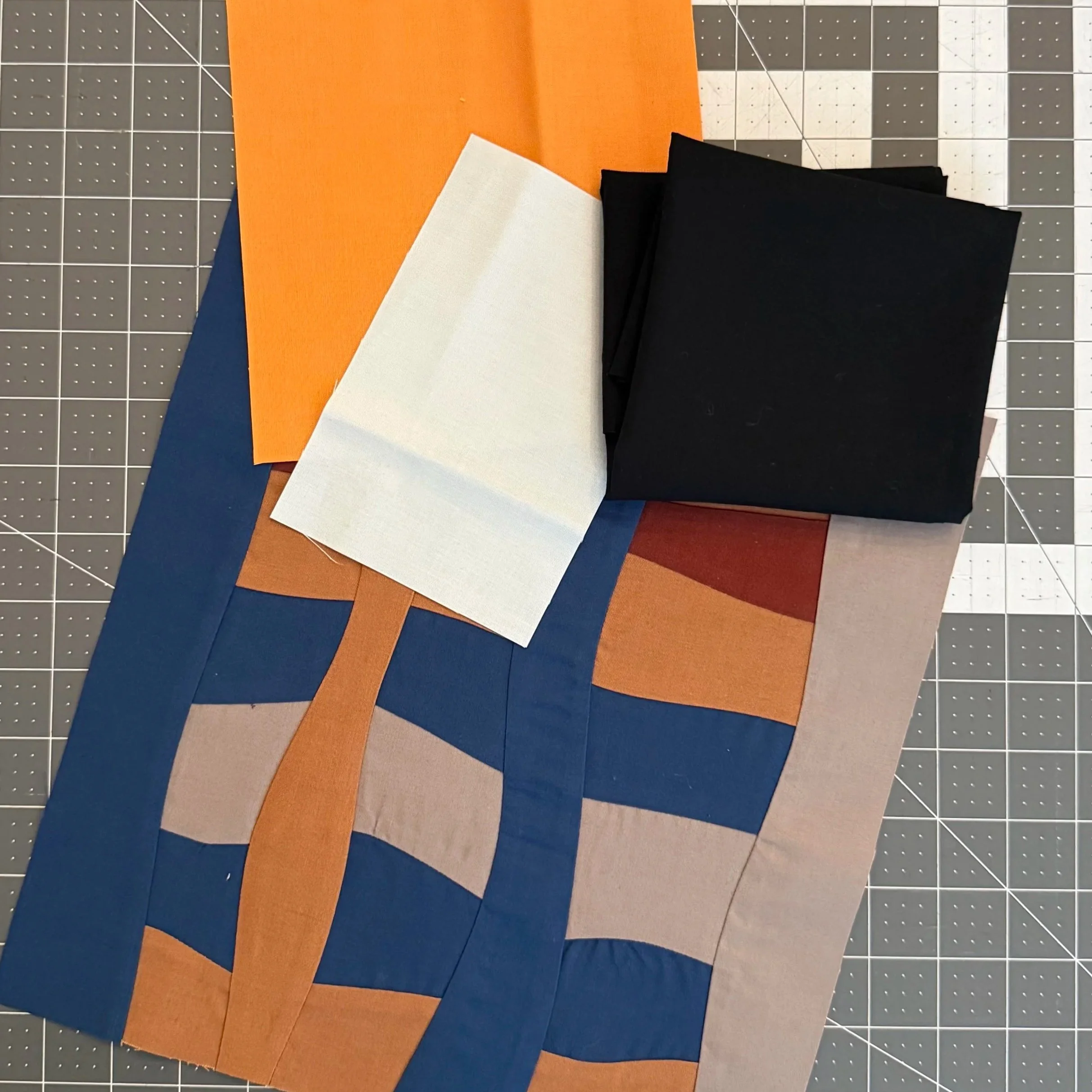 A closeup image of fabric materials in solid colors. One pre-made improvisational quilt block in blue, brown and grey hues is on the bottom resting on a cutting mat. Three pieces of fabric, in orange, ice blue and black rest casually on top.