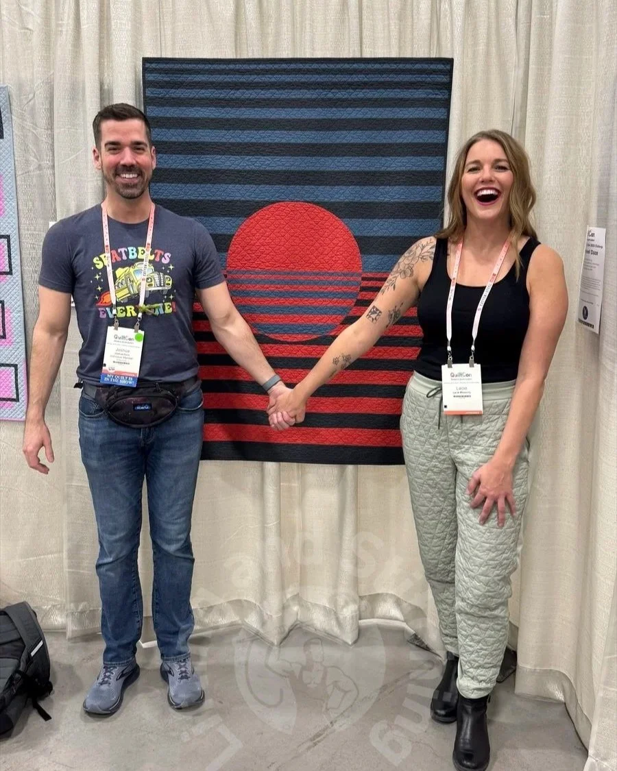 Two quilters hold hands standing in front of a quilt they collaborated together with to make.