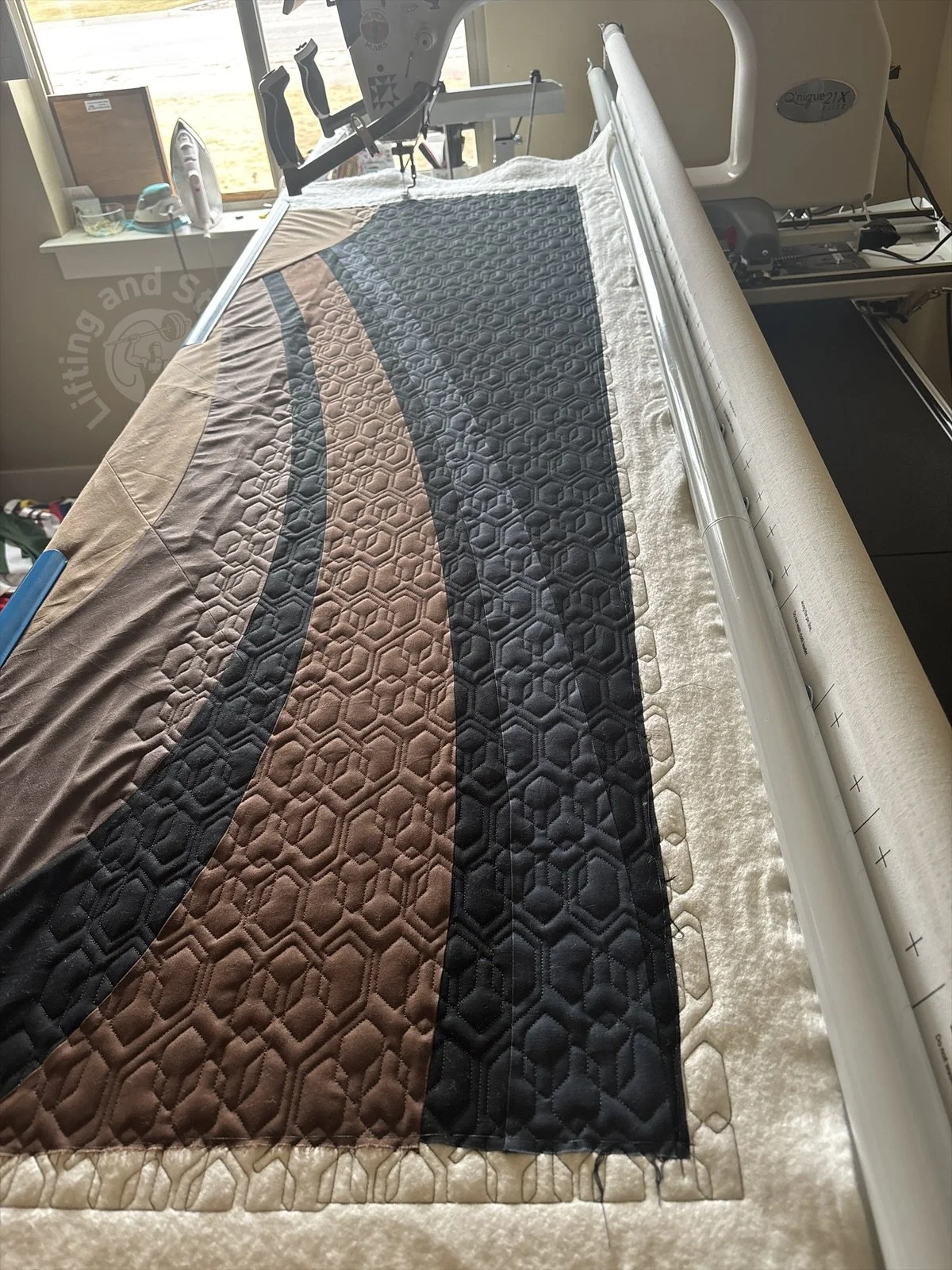 A quilt of Saturn lays on a longarm quilting frame with an edge-to-edge design of hexagons being stitched in a blue-green thread.