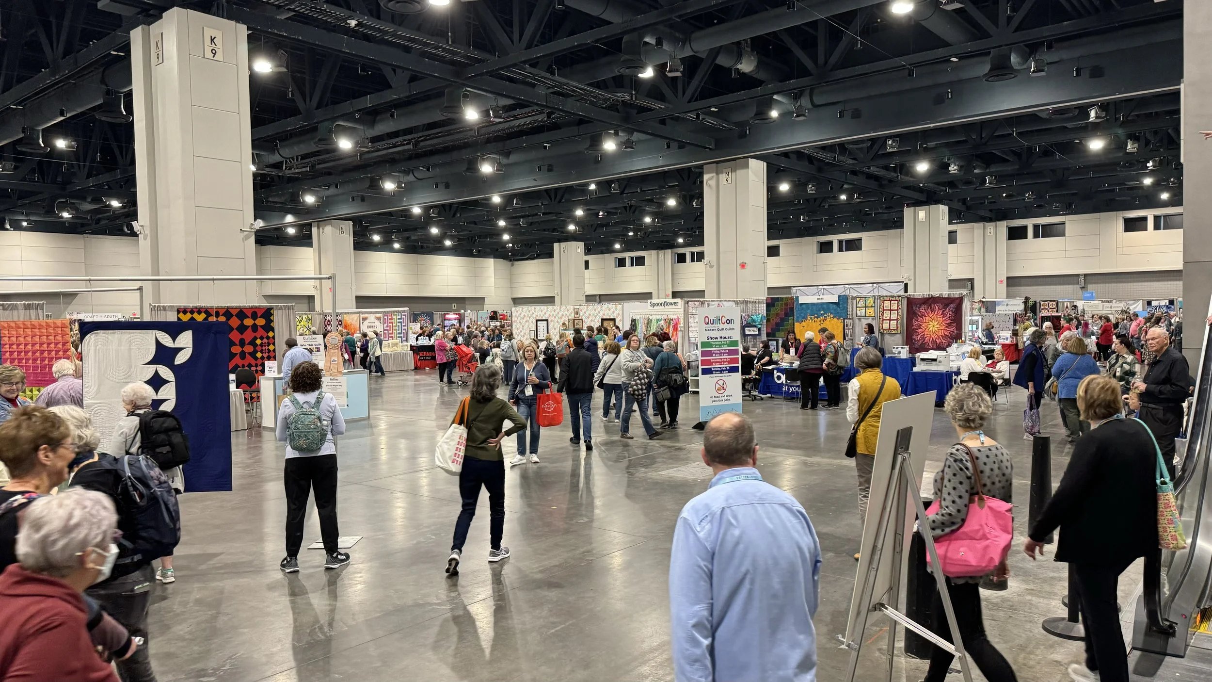 A crowd photo of a quilt show floor. People walking the floor see hundreds of modern quilts hanging on walls.