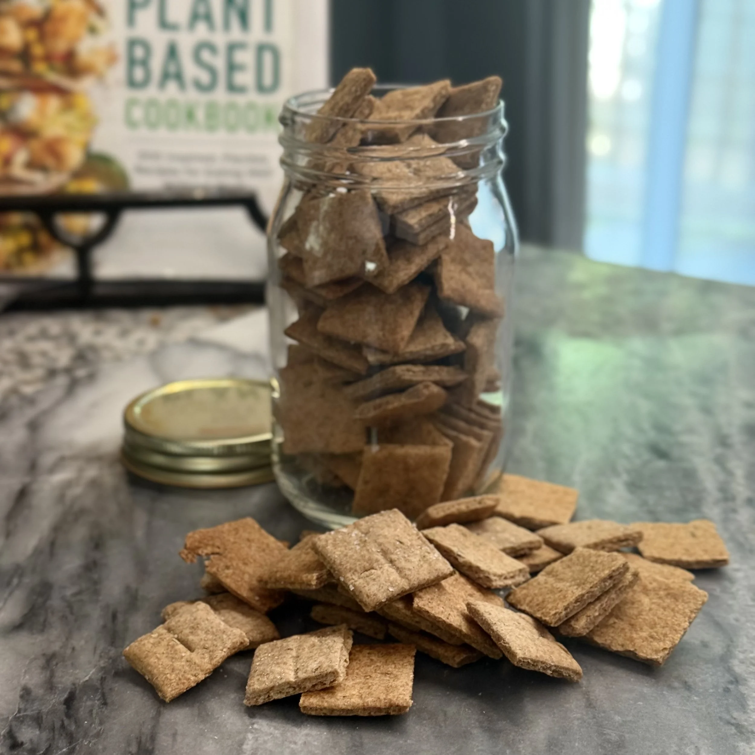 A jar overflowing with homemade crackers. A stack is casually resting on a marble cutting board.