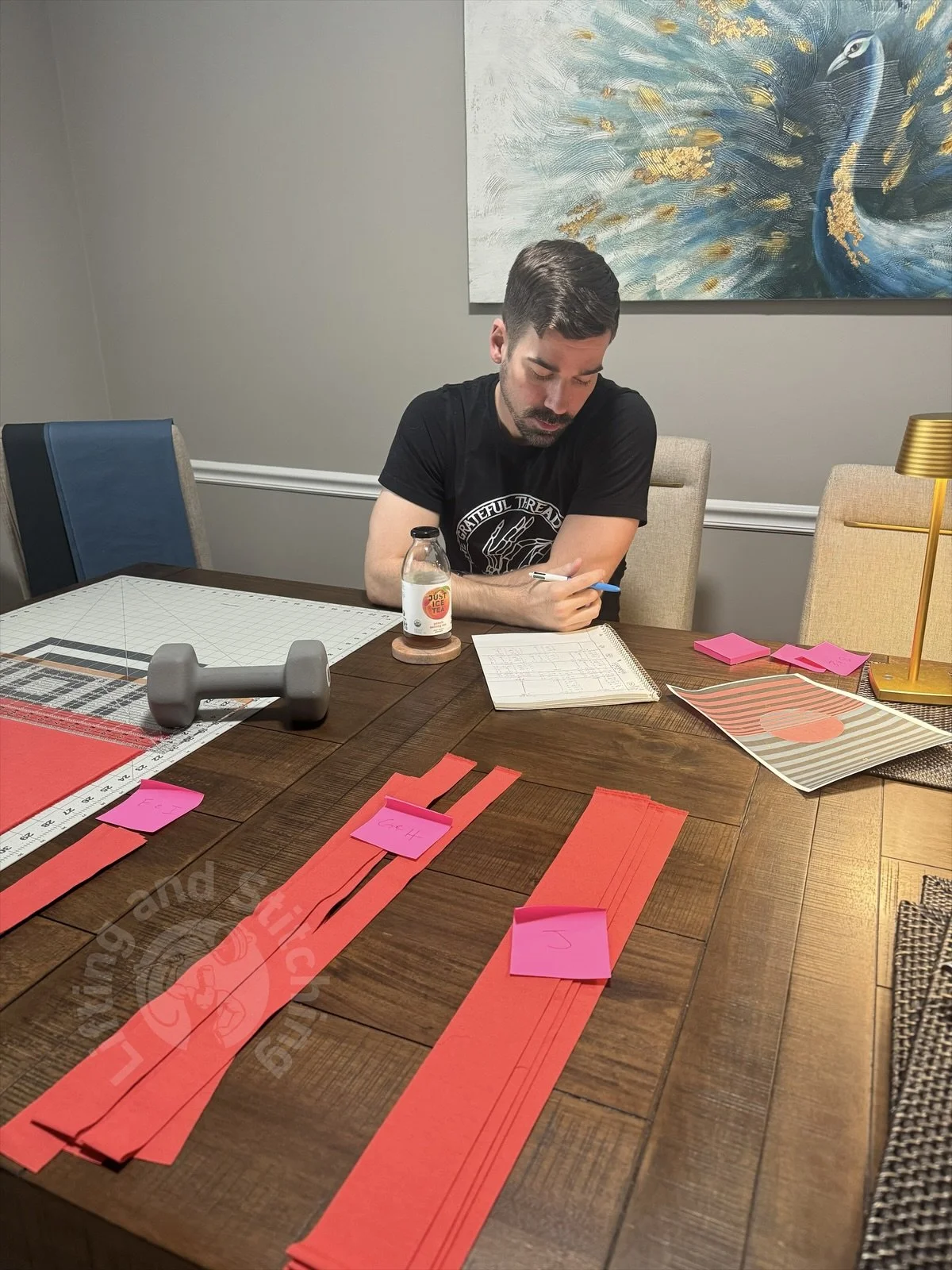 An image of the quilter sitting at a table sketching out and planning an original design. Pieces of cut fabric lay organized across the table in front of him and a cutting board to the left.