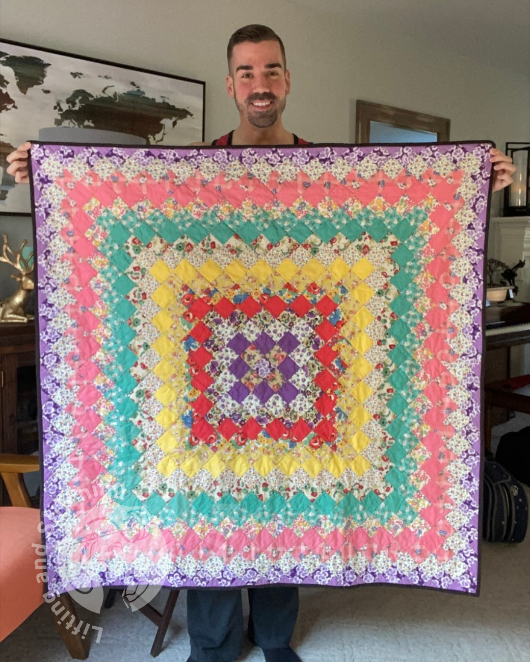 A person stands indoors holding a colorful quilt made of small diamond pieces arranged in concentric squares that shift from purple to red, yellow, green, pink, and a floral border.