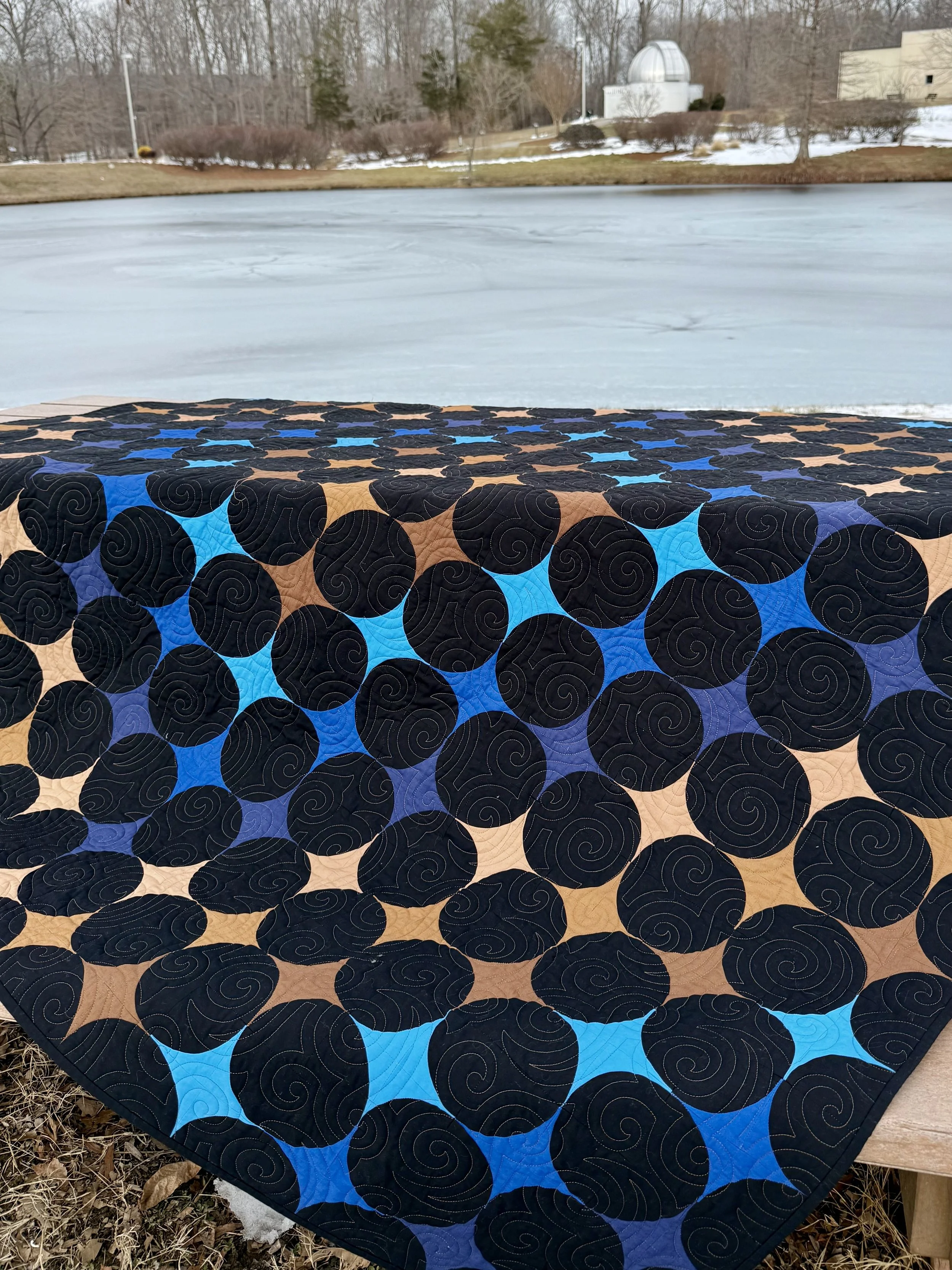 Quilt titled A Trip Around the Universe with a flowing wave of brown and blue fabric arranged into retro-modern four-point stars, by the designer Joshua Dunn of Lifting and Stitching, resting on a picnic table.