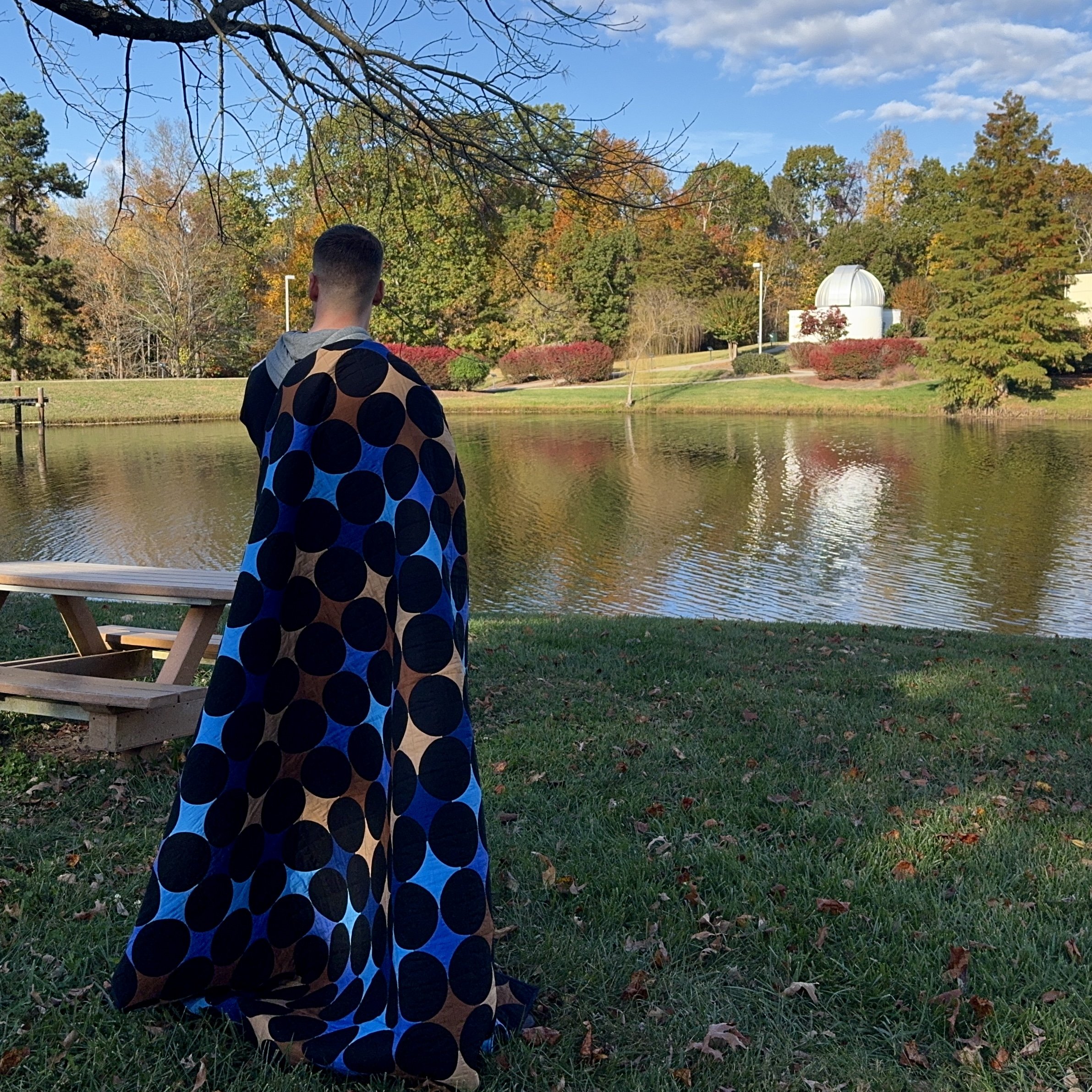 Joshua, a quilter, standing with his back facing the camera with a quilt hanging from his shoulder. He stares at a still pond and off in the distance is an observatory.
