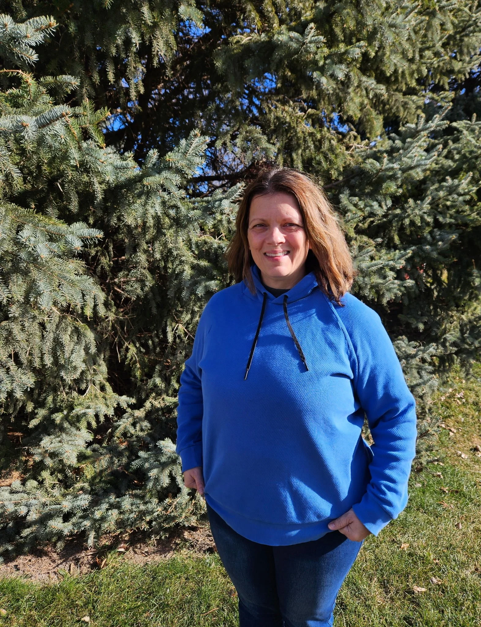 A woman in a blue hoodie standing outdoors in front of a large evergreen tree, smiling at the camera on a sunny day.