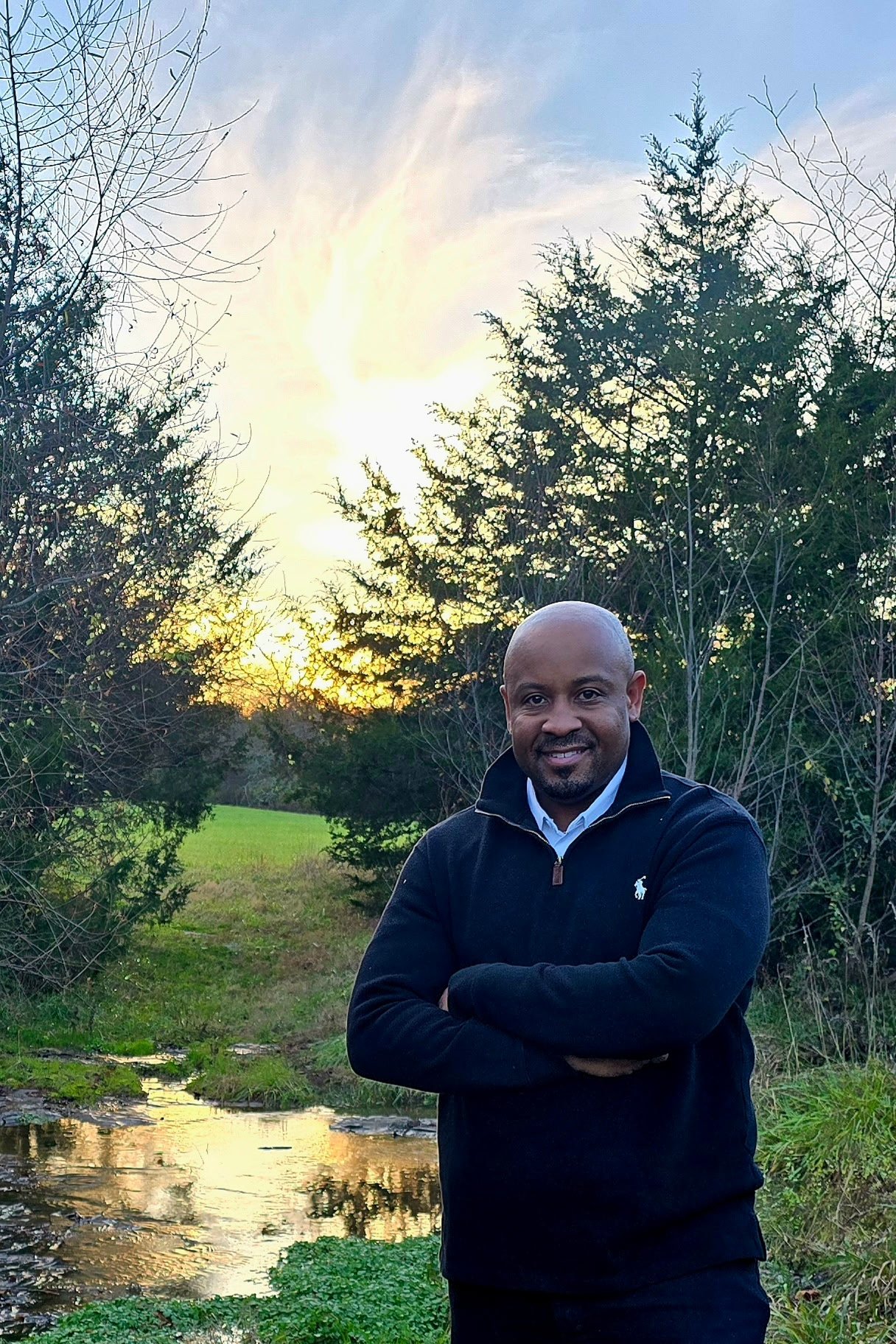 A man in a black sweater standing outdoors near a small creek, with trees and a sunset in the background.