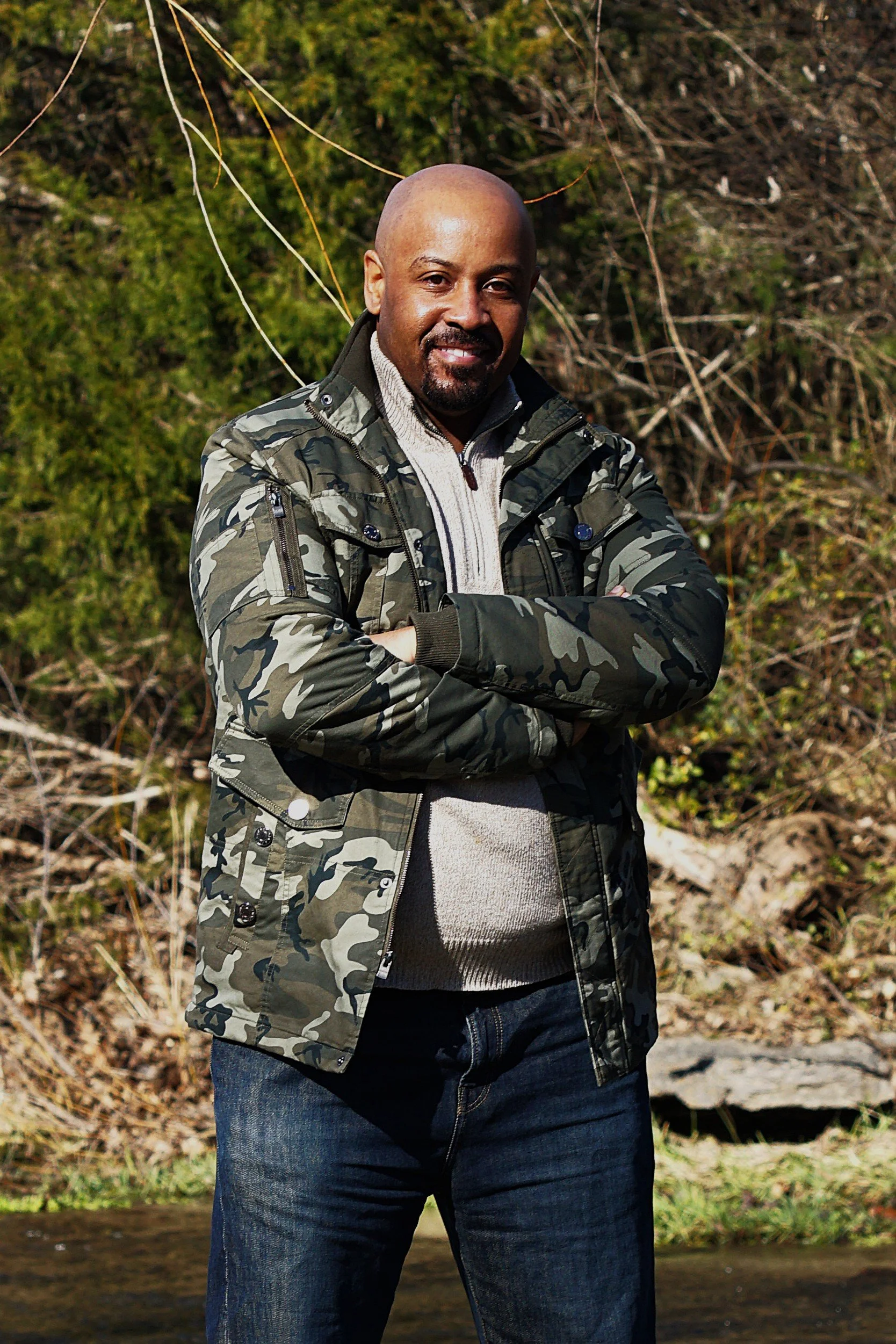 A man in a black sweater standing outdoors near a small creek, with trees and a sunset in the background.