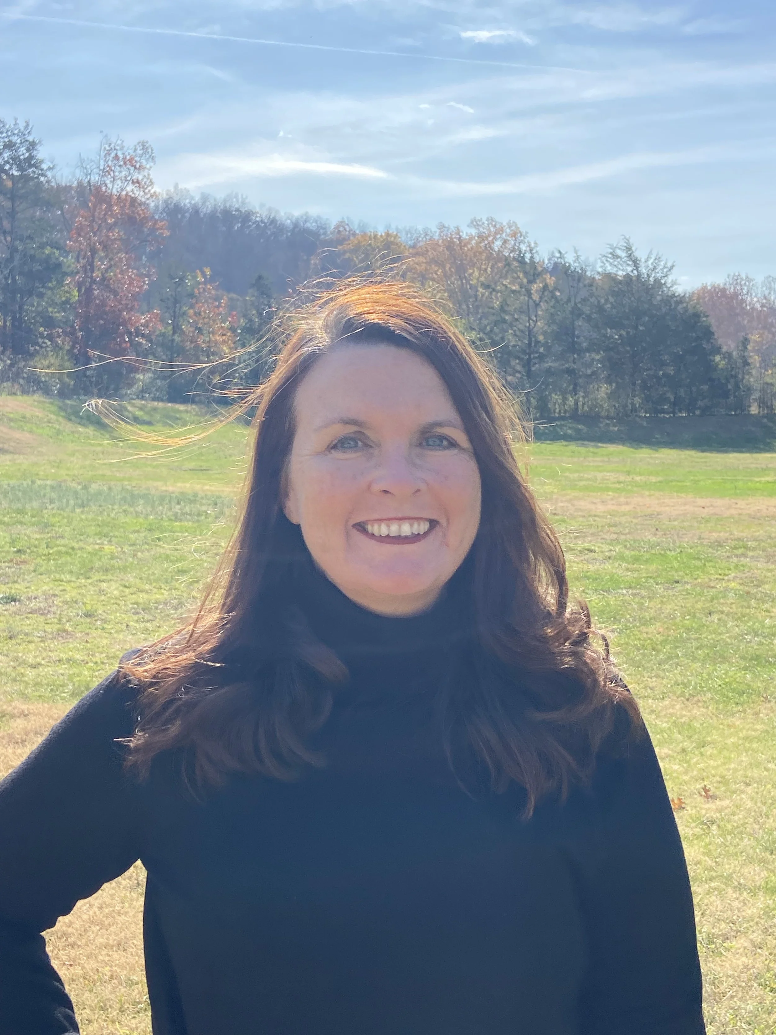 Woman with long brown hair smiling outdoors on a sunny day, wearing a black top, with a grassy field and trees in the background.