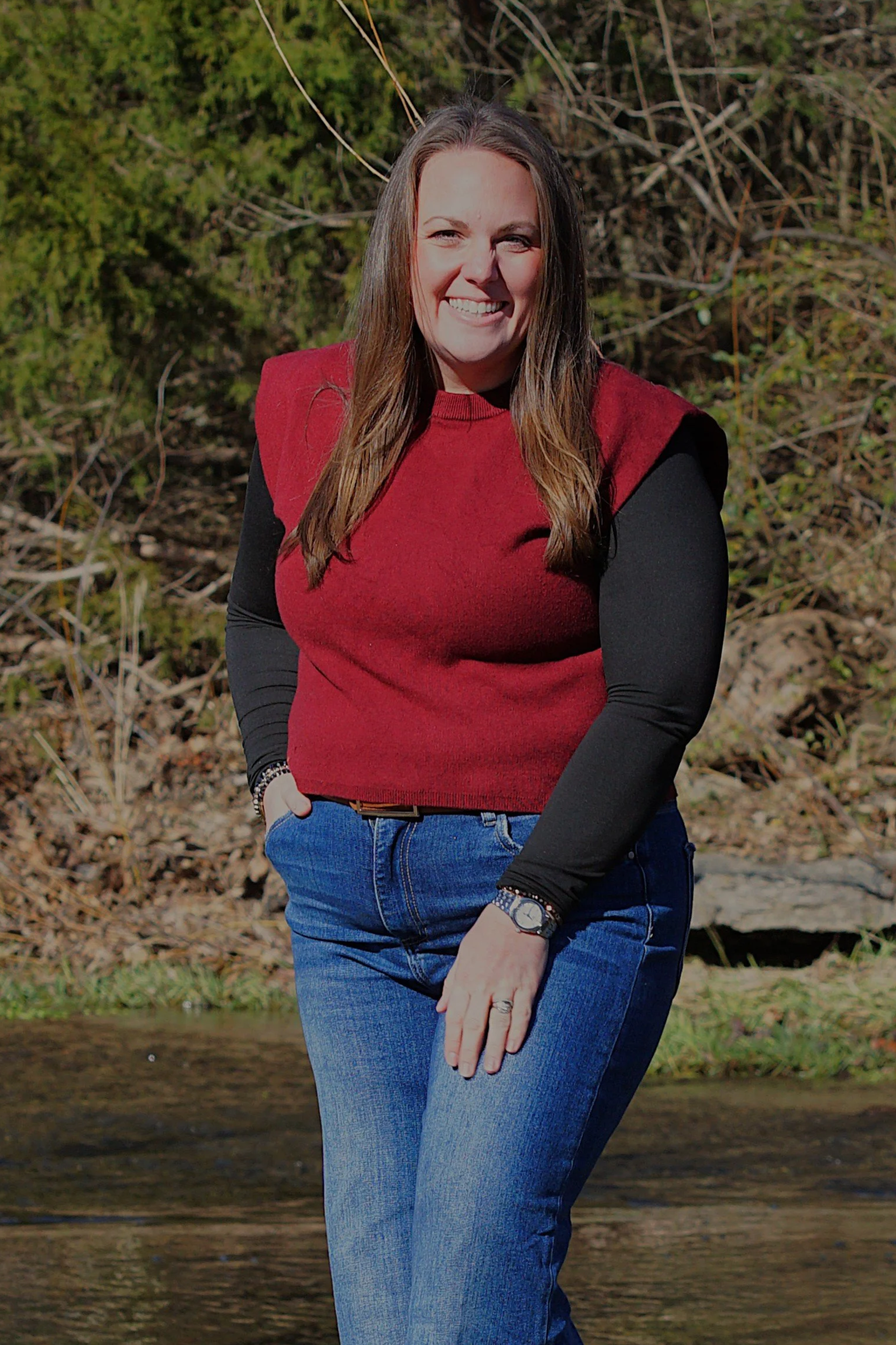 A smiling woman with brown hair wearing a pink dress outdoors with green foliage in the background.