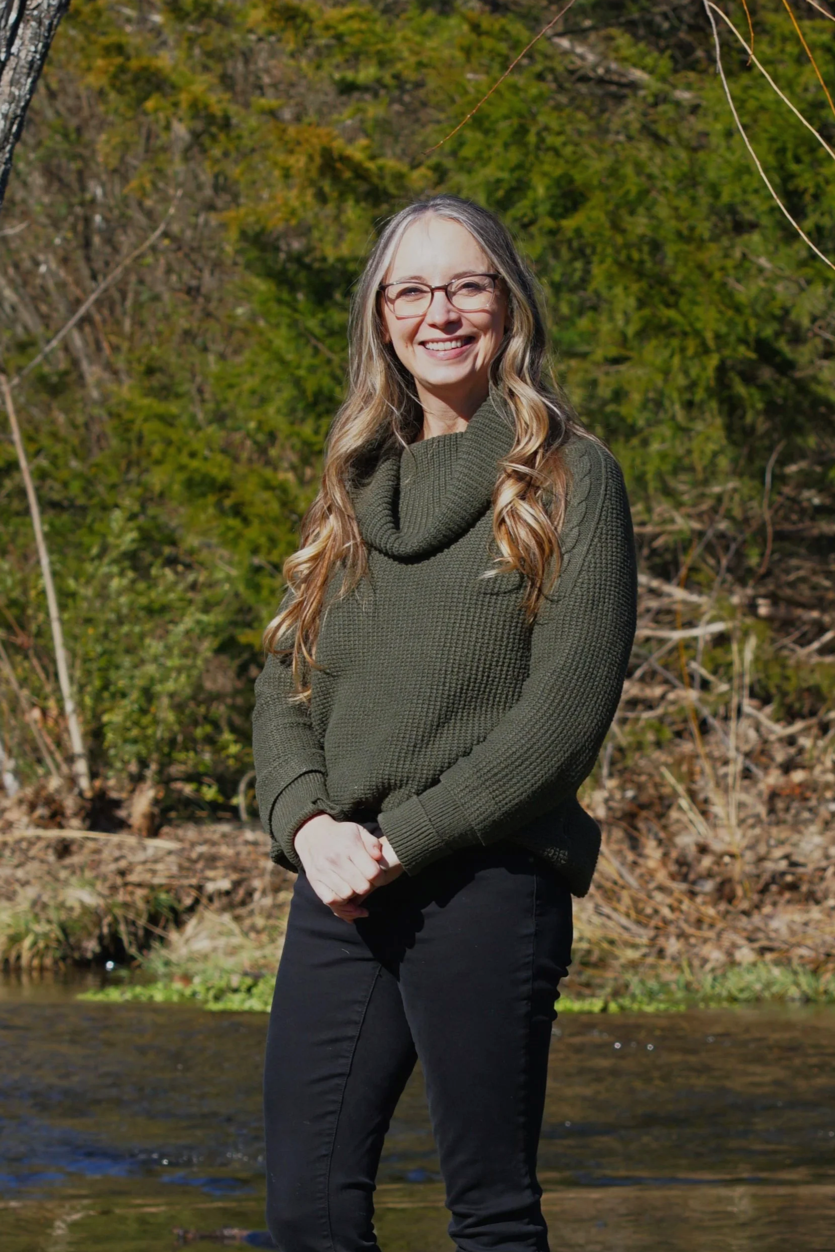 A woman with long gray and brown hair, wearing a red top with buttons and a gold necklace, standing outdoors in front of green foliage and a garden setting.