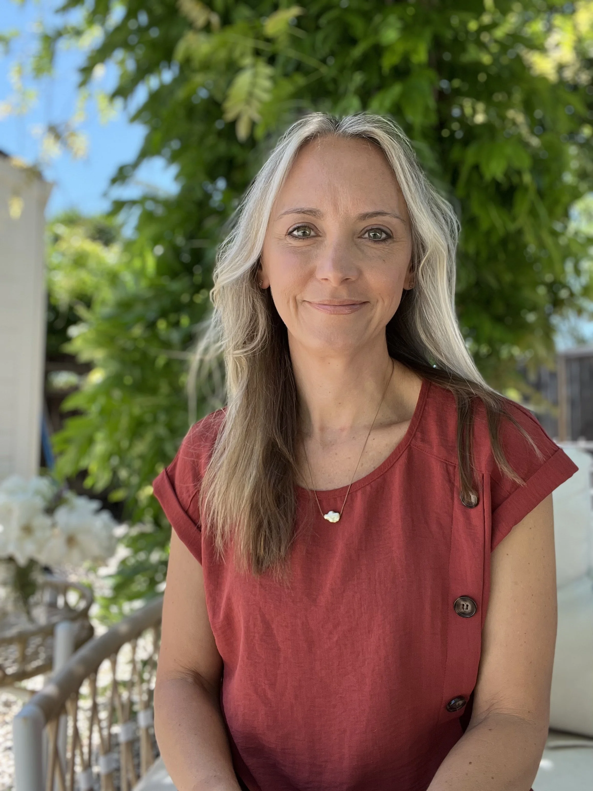 A woman with long gray and brown hair, wearing a red top with buttons and a gold necklace, standing outdoors in front of green foliage and a garden setting.