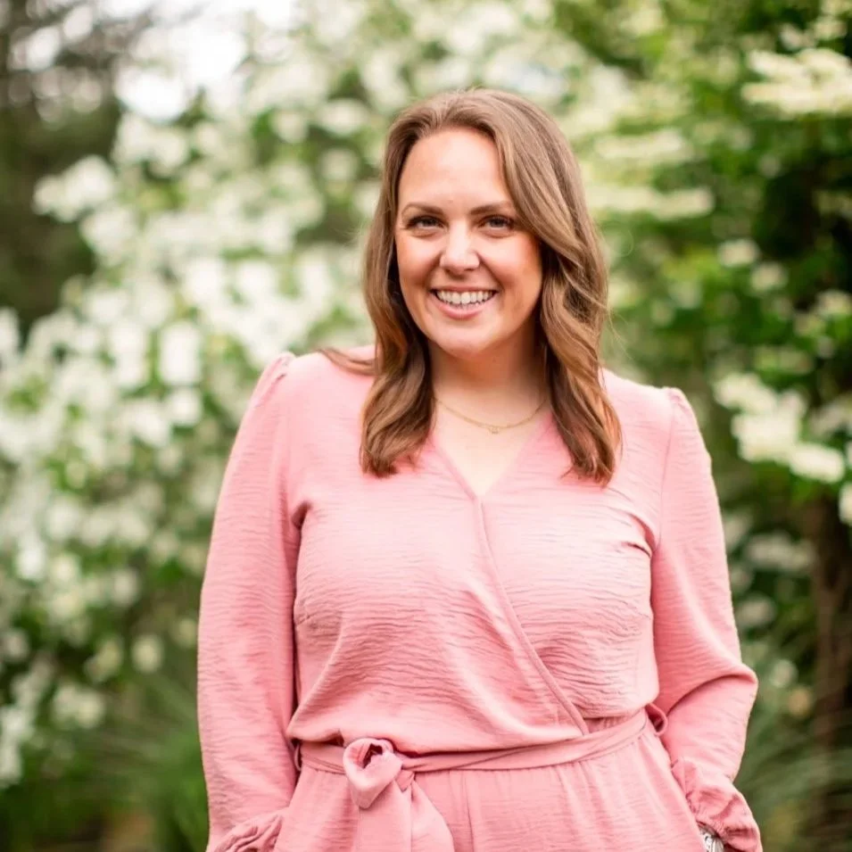 A smiling woman with brown hair wearing a pink dress outdoors with green foliage in the background.