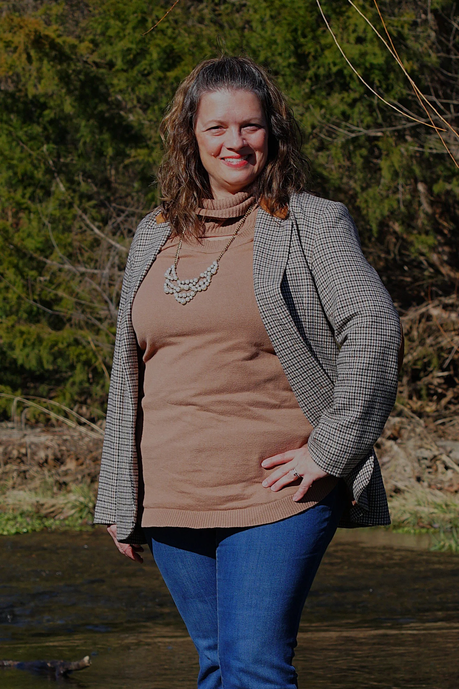 A woman in a blue hoodie standing outdoors in front of a large evergreen tree, smiling at the camera on a sunny day.
