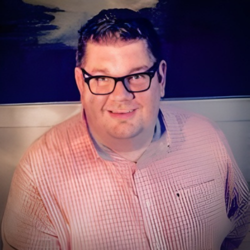 A man with dark hair, glasses, and a light pink checkered shirt smiling at the camera, standing outdoors at night with a body of water and the moon in the background.
