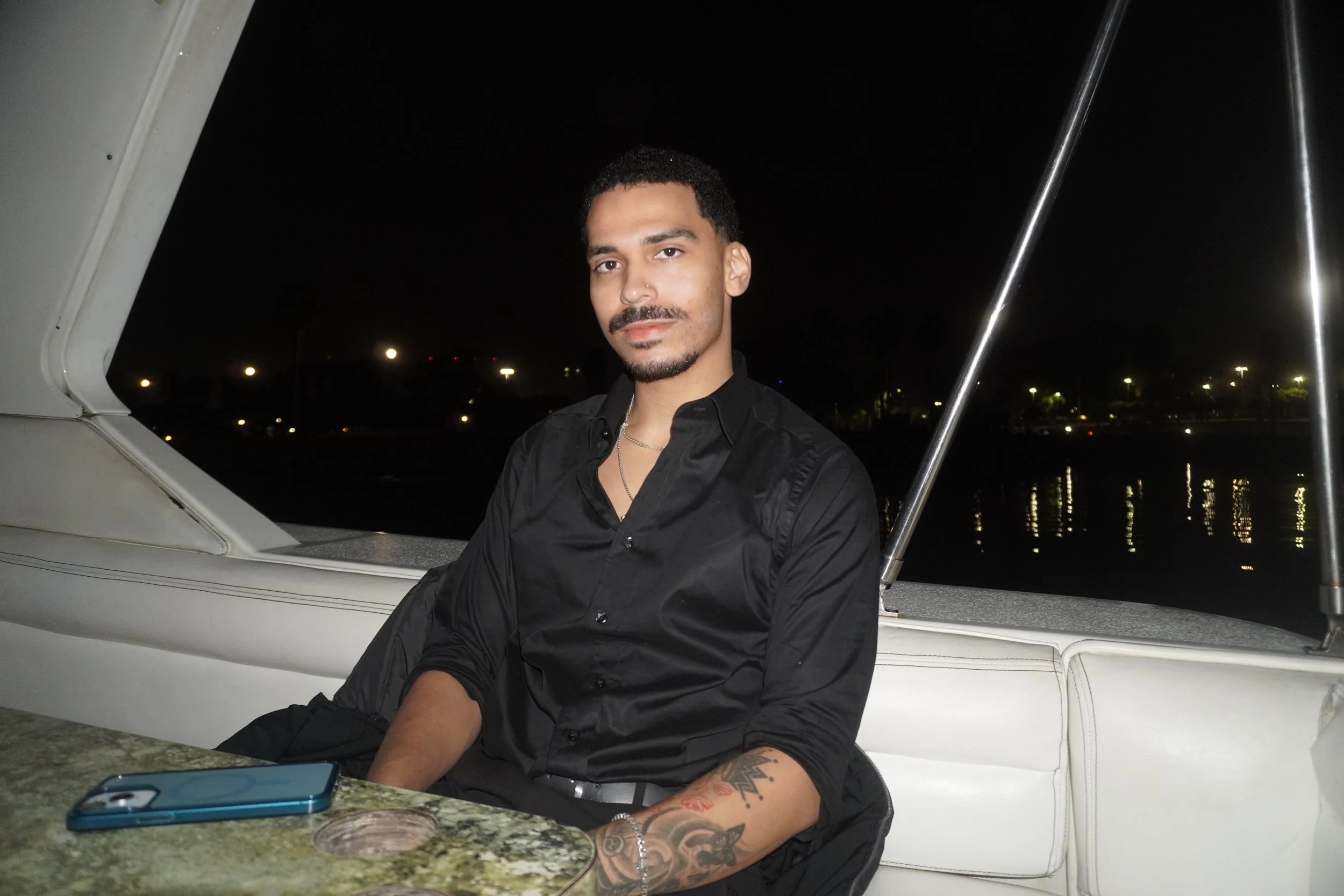 A young man with dark hair, a mustache, and tattoos on his left arm, wearing a black shirt, sitting on a boat at night with city lights reflecting on the water in the background.