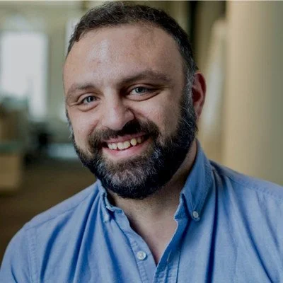 Close-up of smiling man with dark hair and beard wearing a blue button-up shirt