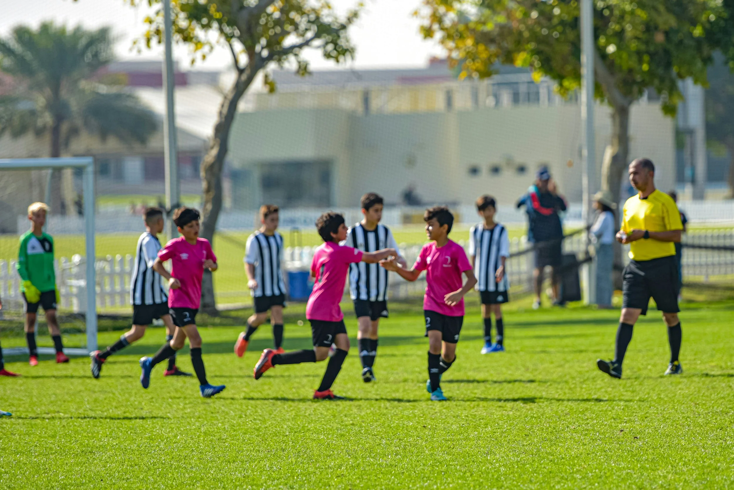 Young soccer players in pink and black uniforms celebrating after scoring a goal during a match on a sunny day with a referee and opposing team in black and white stripes.