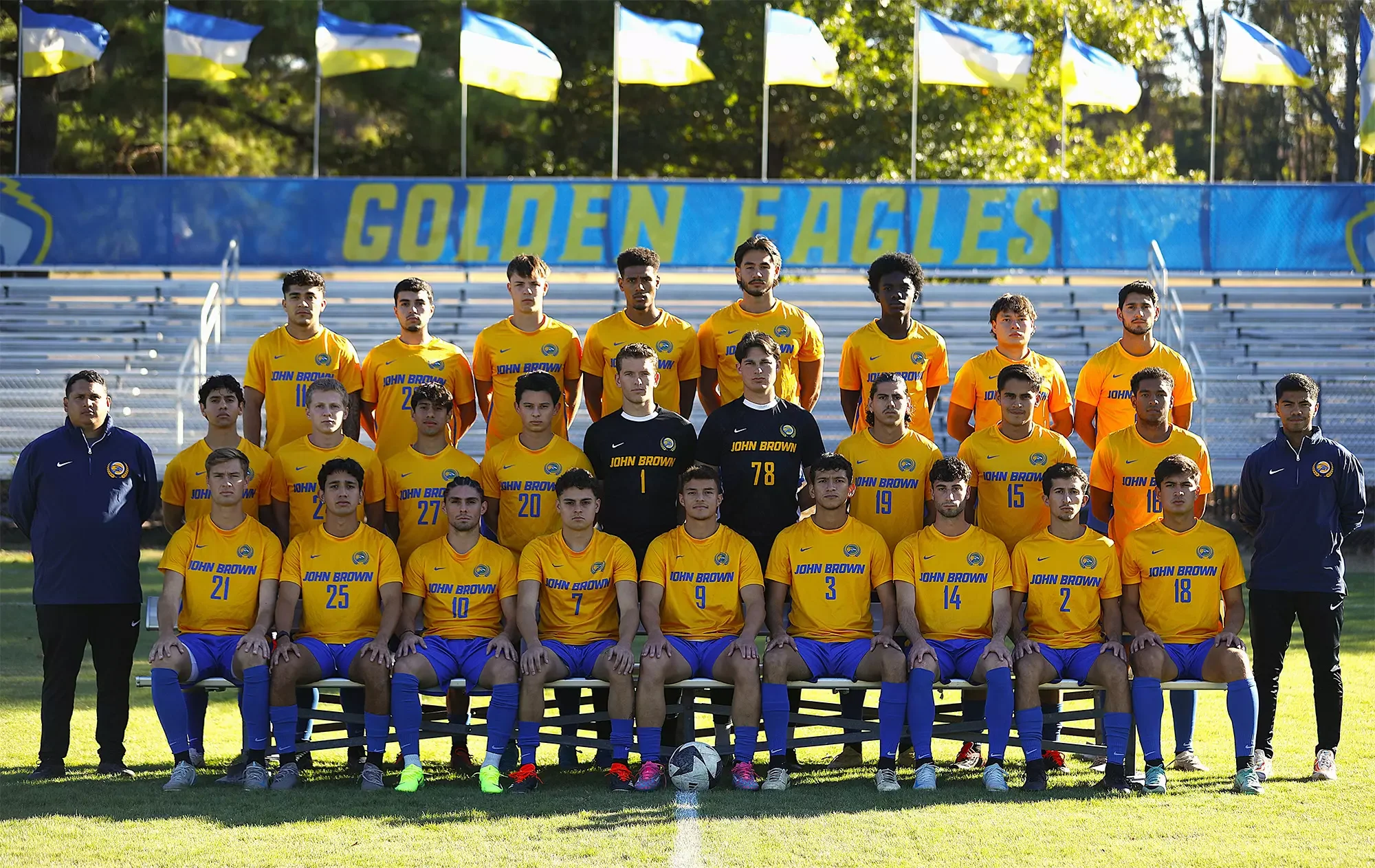 A soccer team in yellow and blue uniforms posing for a group photo on a field, with some players sitting and some standing. There are two coaches wearing navy jackets on either side of the group. In the background, a banner reads 'GOLDEN EAGLES' and there are flags flying.