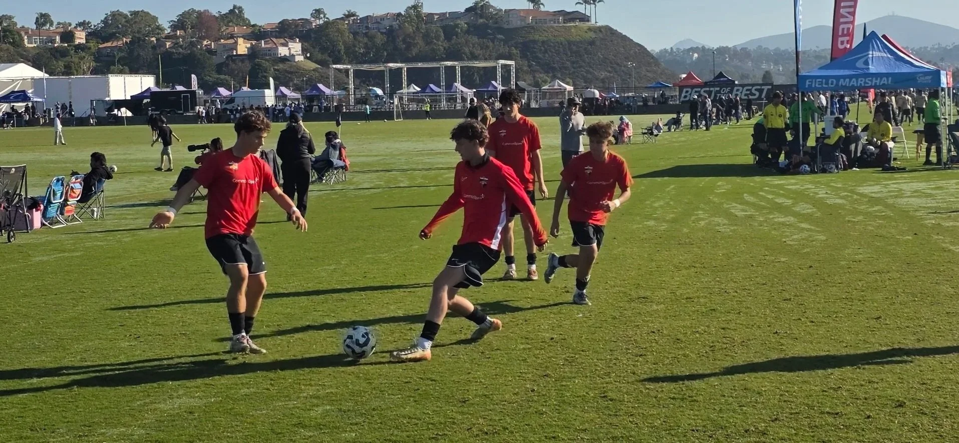 Four boys playing soccer on a grassy field during a sporting event, with tents and spectators in the background.