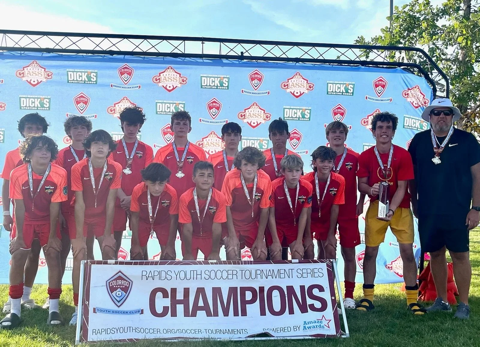 Youth soccer team wearing red and orange jerseys and medals poses for a group photo outdoors, with a coach on the right. They are holding a large banner that reads "CHAMPIONS" at a youth soccer tournament, with a backdrop featuring various sponsor logos.