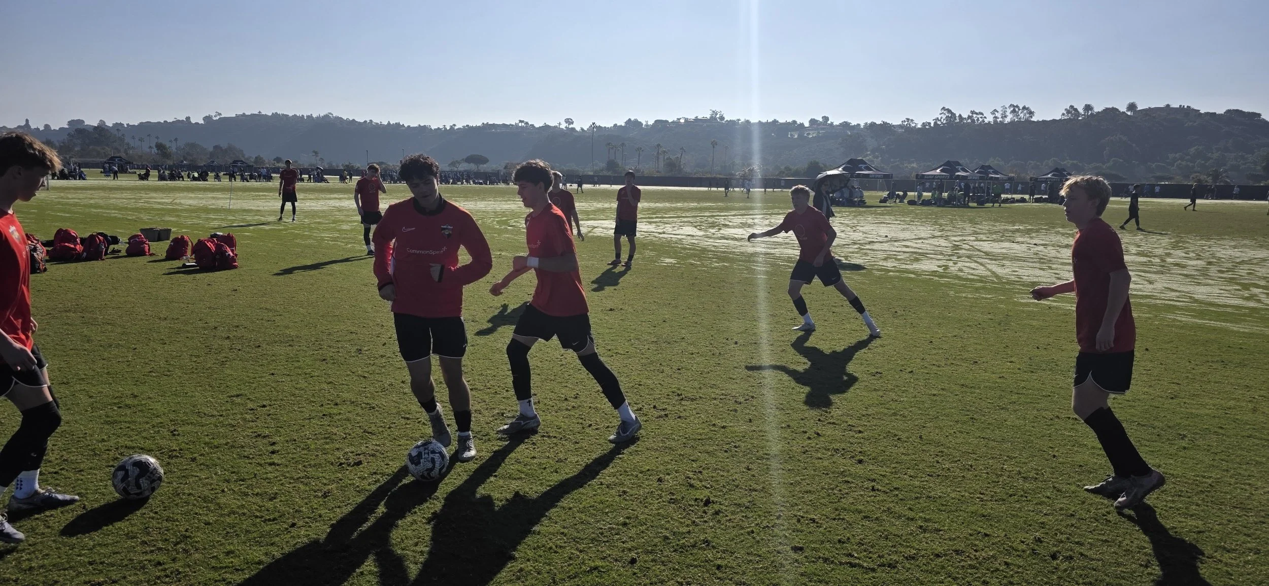 Young soccer players in red shirts practicing on a grassy field with shadows cast on the ground, tents and distant hills in the background.