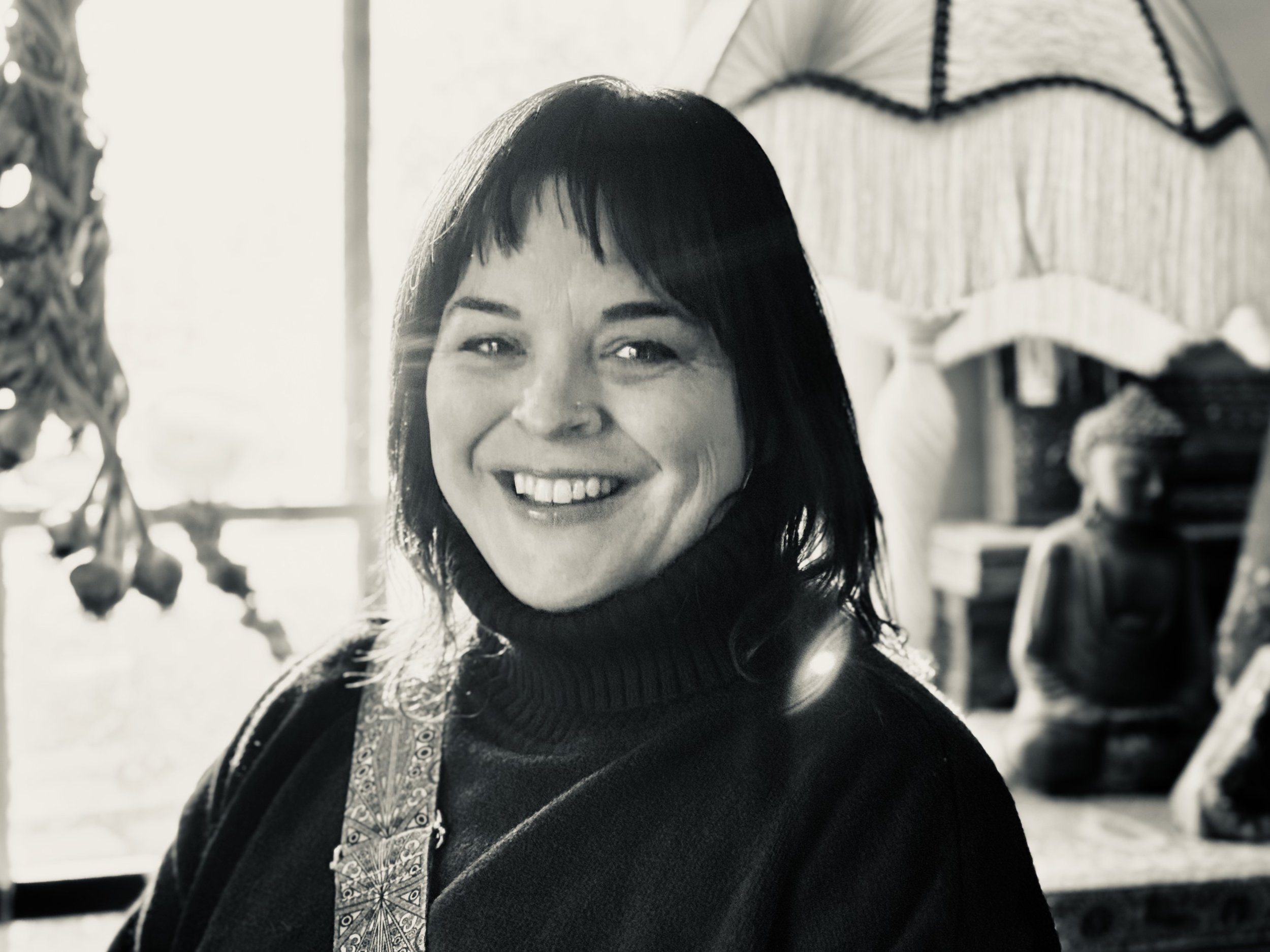 Black-and-white photo of a smiling female doula with dark hair, wearing a turtleneck sweater, indoors with a window and decorative objects in the background.