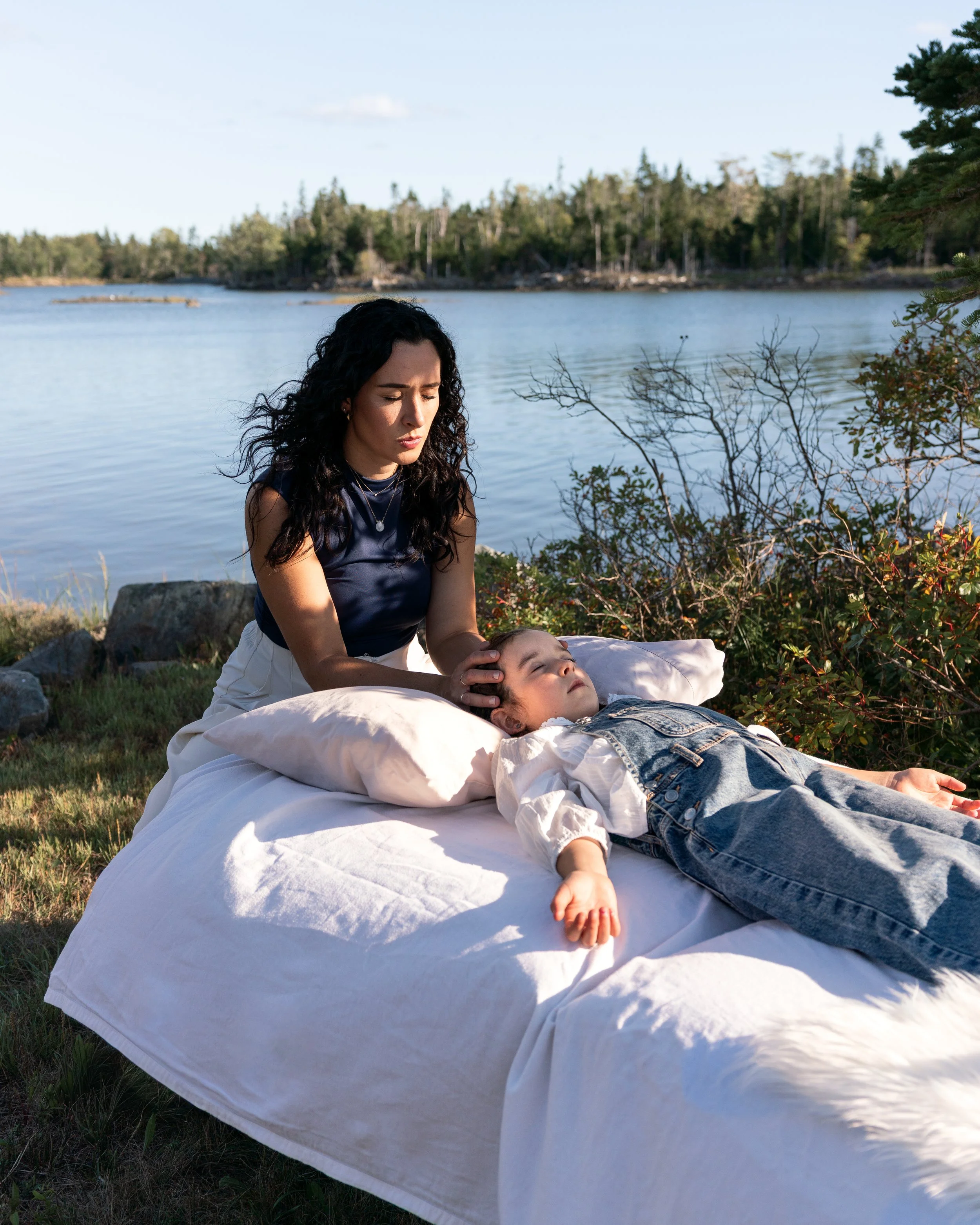 A woman is assessing cranial vitality of a young girl lying on a bed outside near a body of water, with trees in the background.