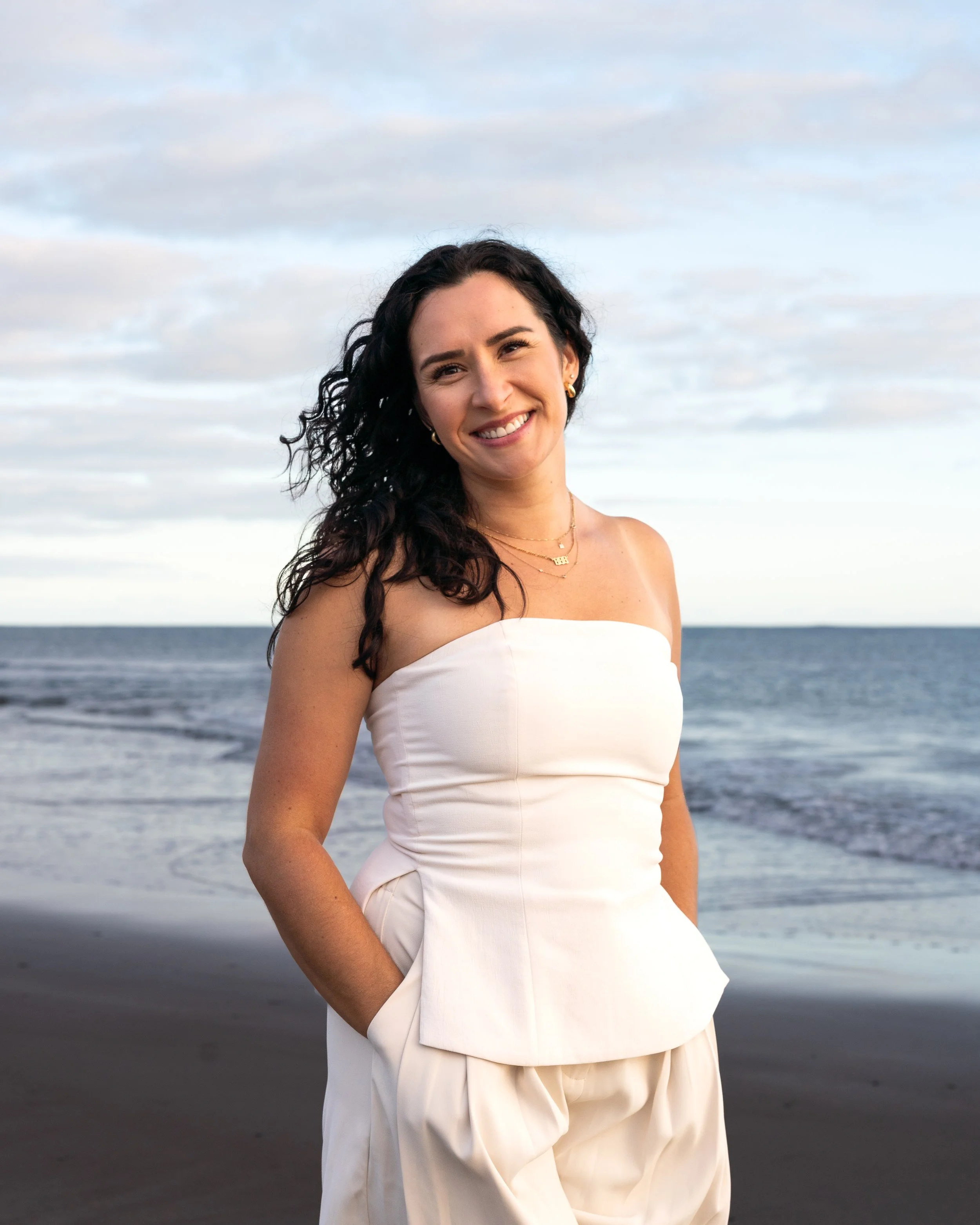 A woman with curly dark hair in a white strapless dress standing on the beach during sunset, with the ocean and cloudy sky in the background.