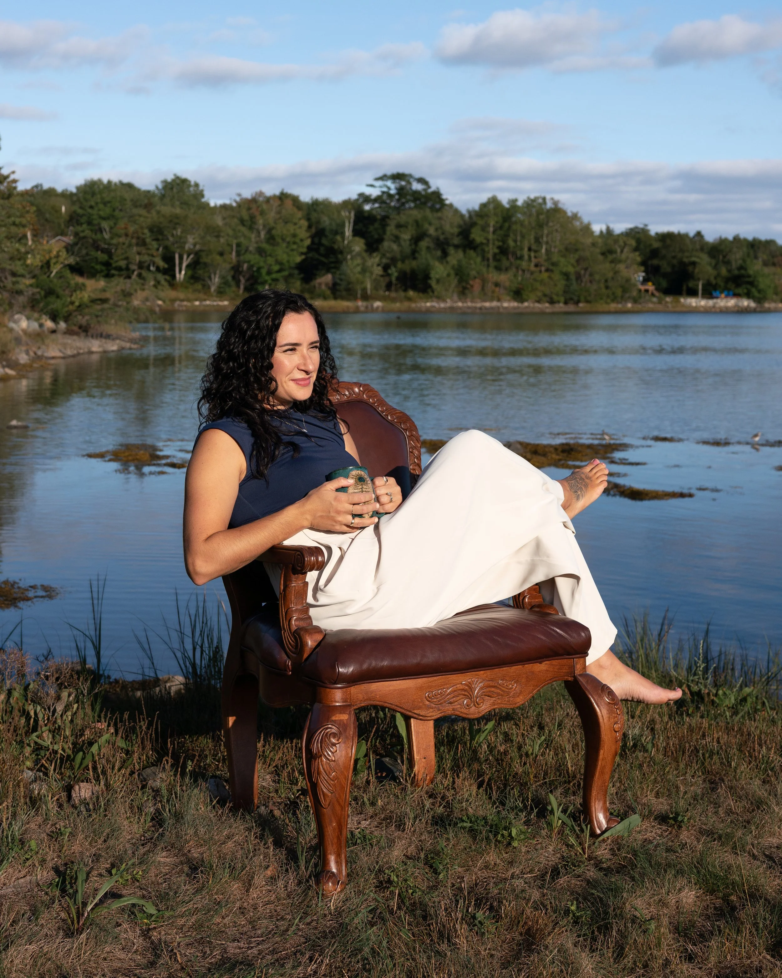 A woman sitting on a wooden chair outdoors by a body of water, holding a mug, with trees and a cloudy sky in the background.