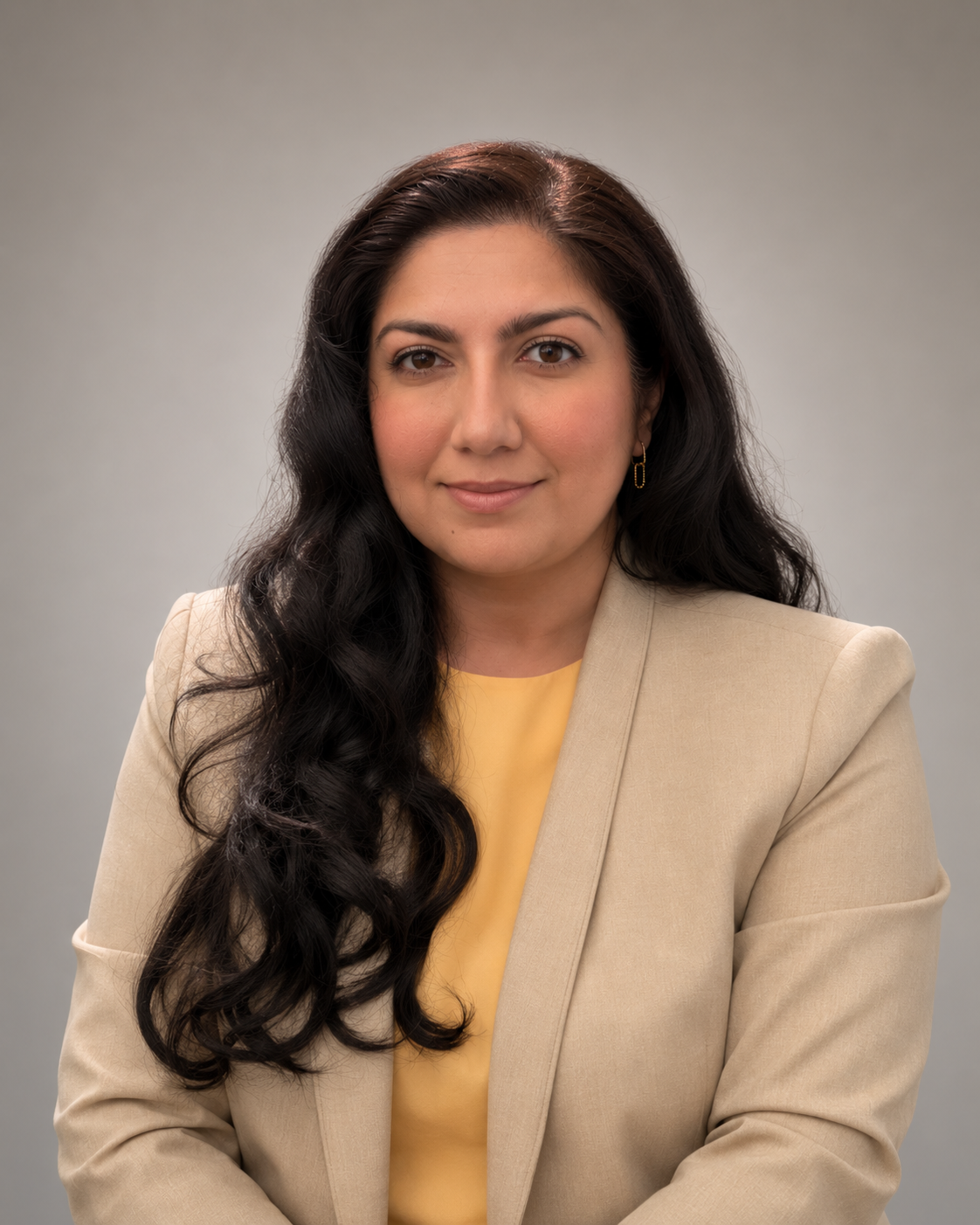 A woman with long, dark, wavy hair wearing a beige blazer and yellow top, posed against a plain gray background.