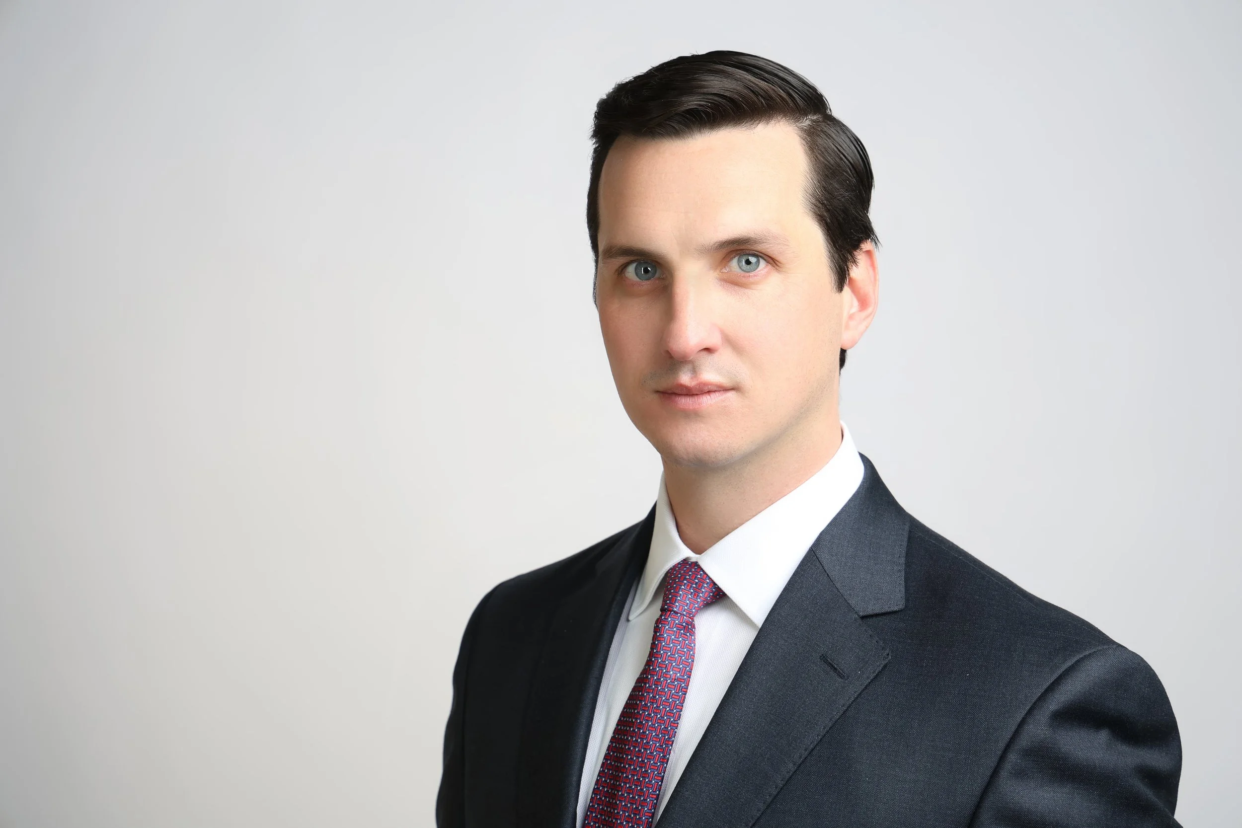 Professional portrait of a man in a dark suit, white shirt, and red tie, standing against a plain light gray background.