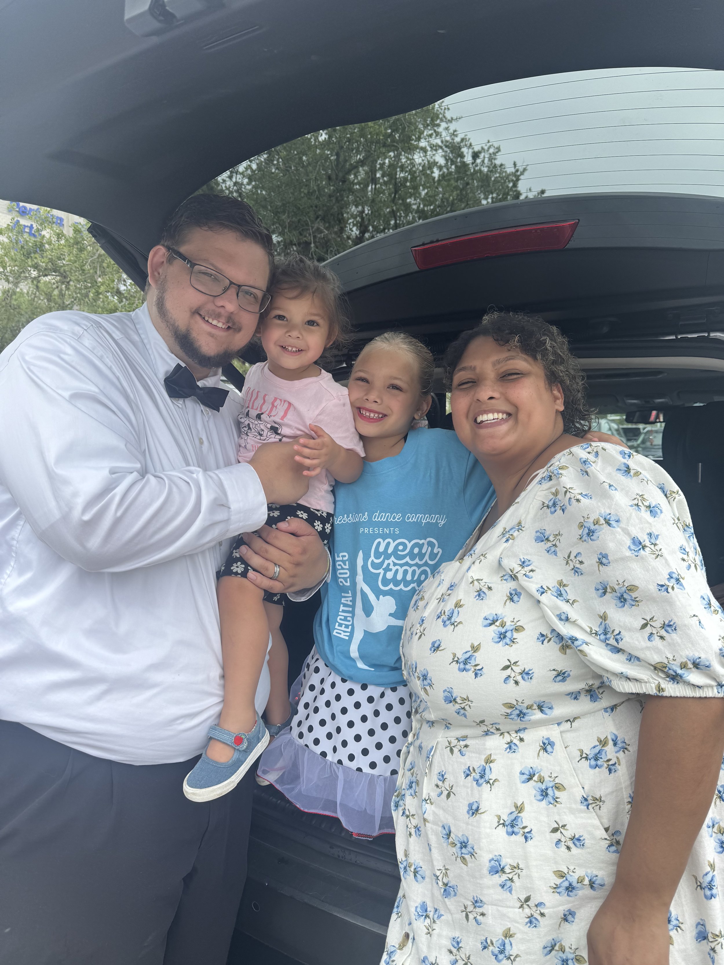 Family of four smiling and posing together near a car trunk in an outdoor setting.