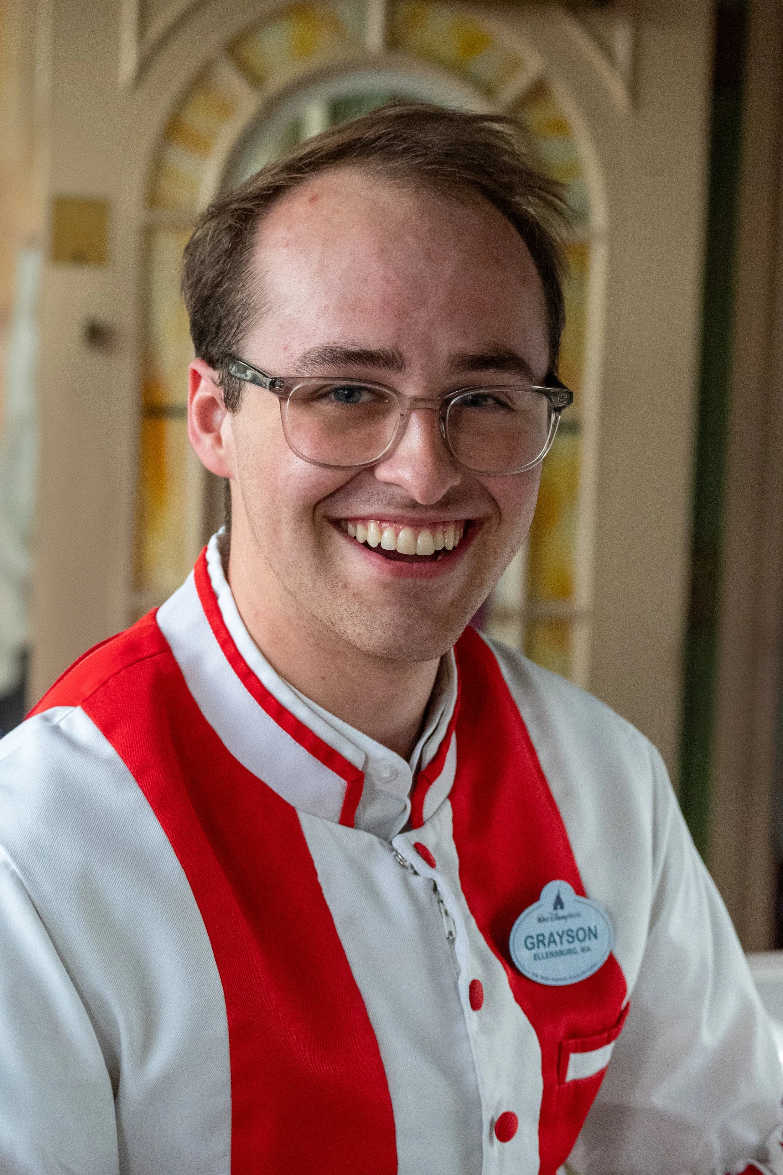 A young man with glasses, short brown hair, and a bright smile, wearing a white and red uniform with a name tag that reads 'Grayson', standing indoors in front of a stained glass window.