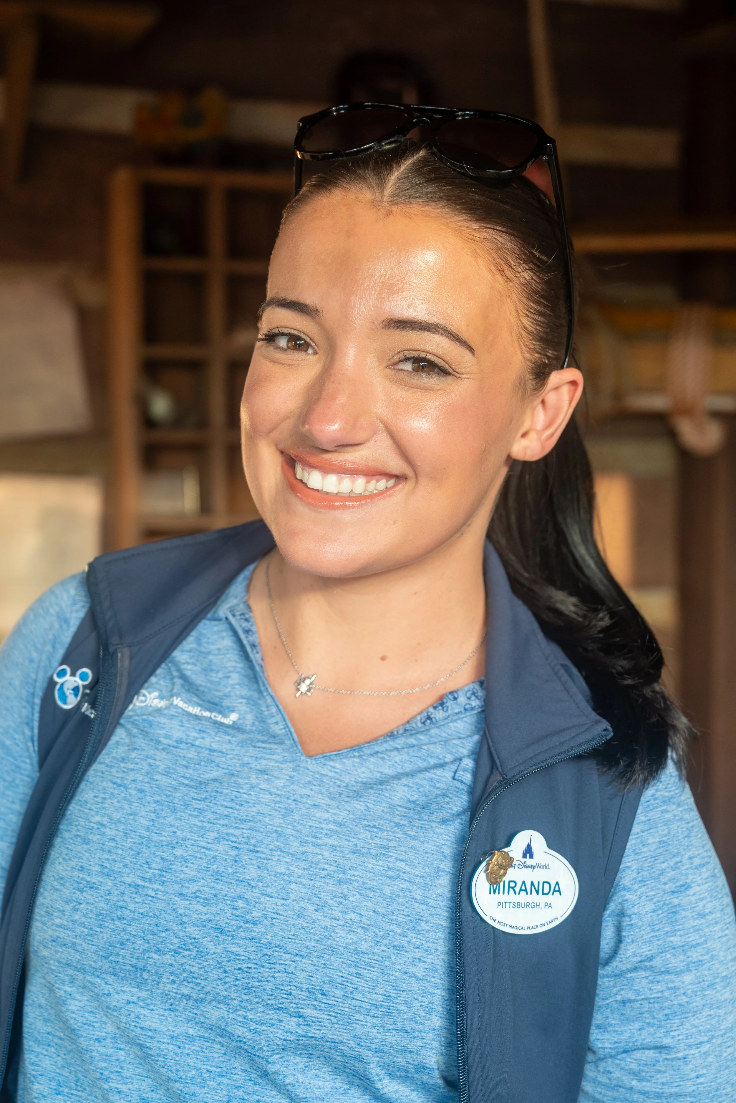 Smiling woman with sunglasses on her head, wearing a blue shirt and a vest with a name tag that says 'Miranda' in Pittsburgh, PA, indoors.