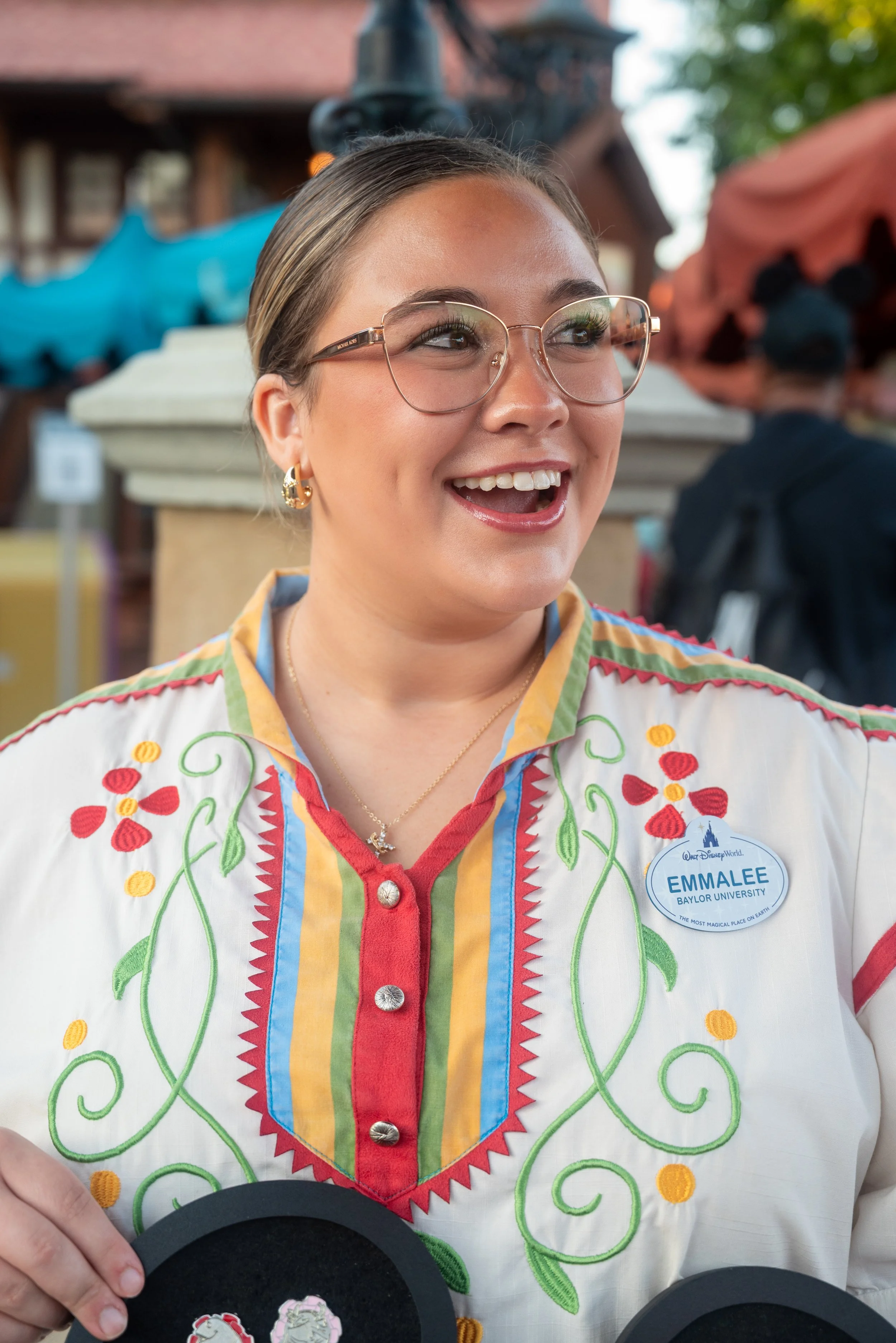 A woman with glasses and earrings, smiling, wearing a colorful embroidered shirt with a Dallas Disney World badge that says 'Emalee Baylor University,' standing outdoors at a festive event.