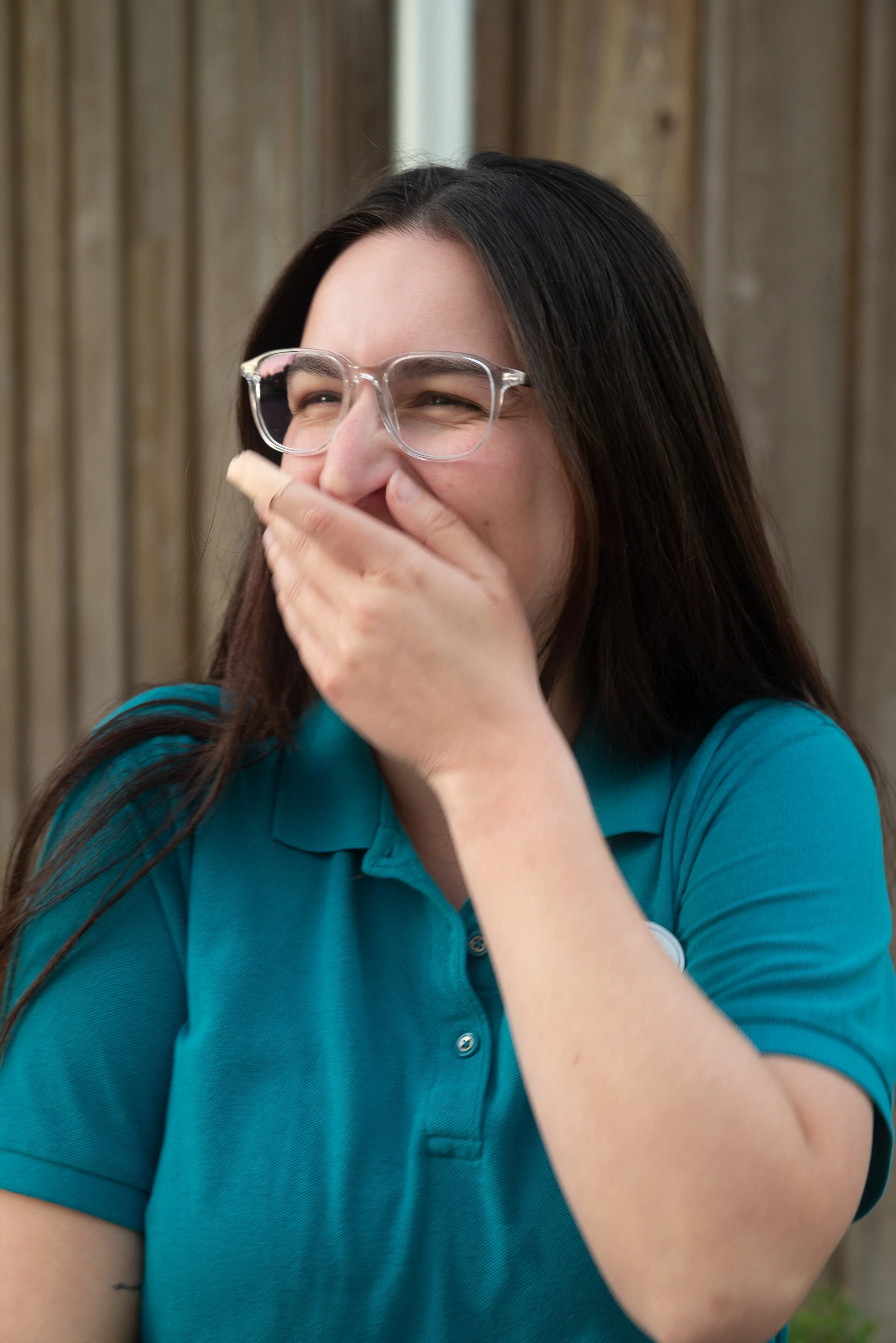 A woman with long dark hair and glasses, wearing a teal polo shirt, is smiling and covering her mouth with her hand, standing outdoors with a wooden fence in the background.