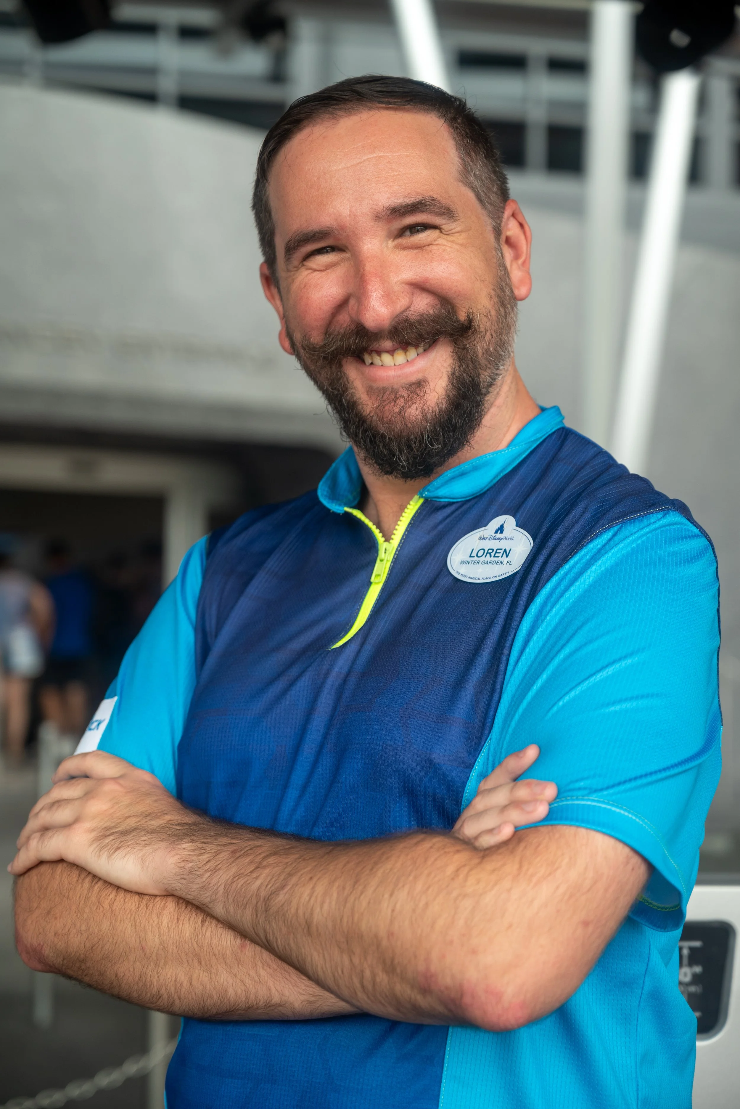 A smiling man with a beard, wearing a blue and teal uniform with a name tag that says 'Loren' from Winter Garden, FL, standing arms crossed indoors.